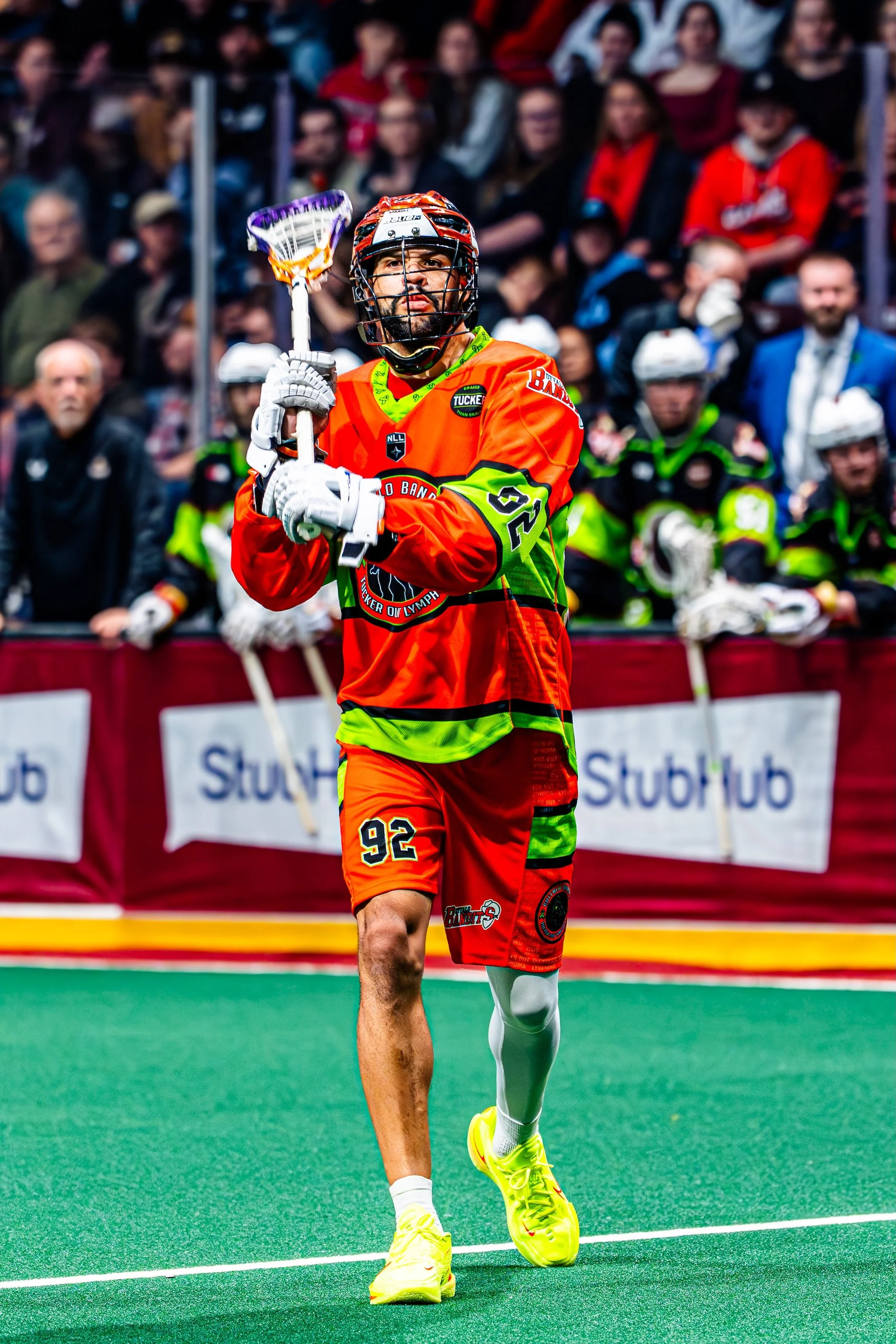 A man in a vibrant orange and green hockey uniform with the number 92 runs on an indoor field. He wears a helmet with a full face cage, holds a hockey stick, and has a focused expression. There are spectators and other players sitting behind a barrie