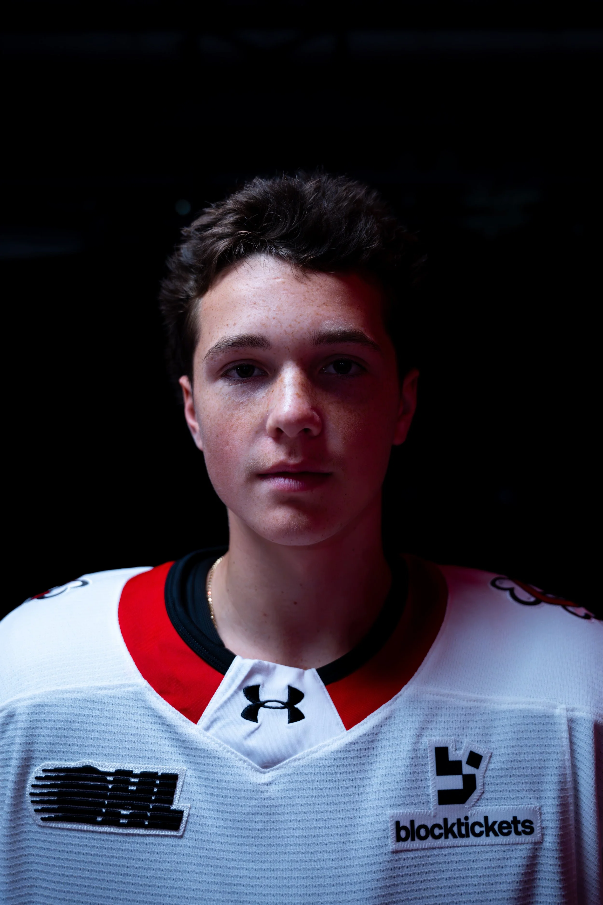 Close-up portrait of a young male athlete with short, dark curly hair, wearing a white sports jersey with red and black accents, and multiple sponsor logos including "blocktickets" and Under Armour, against a black background.