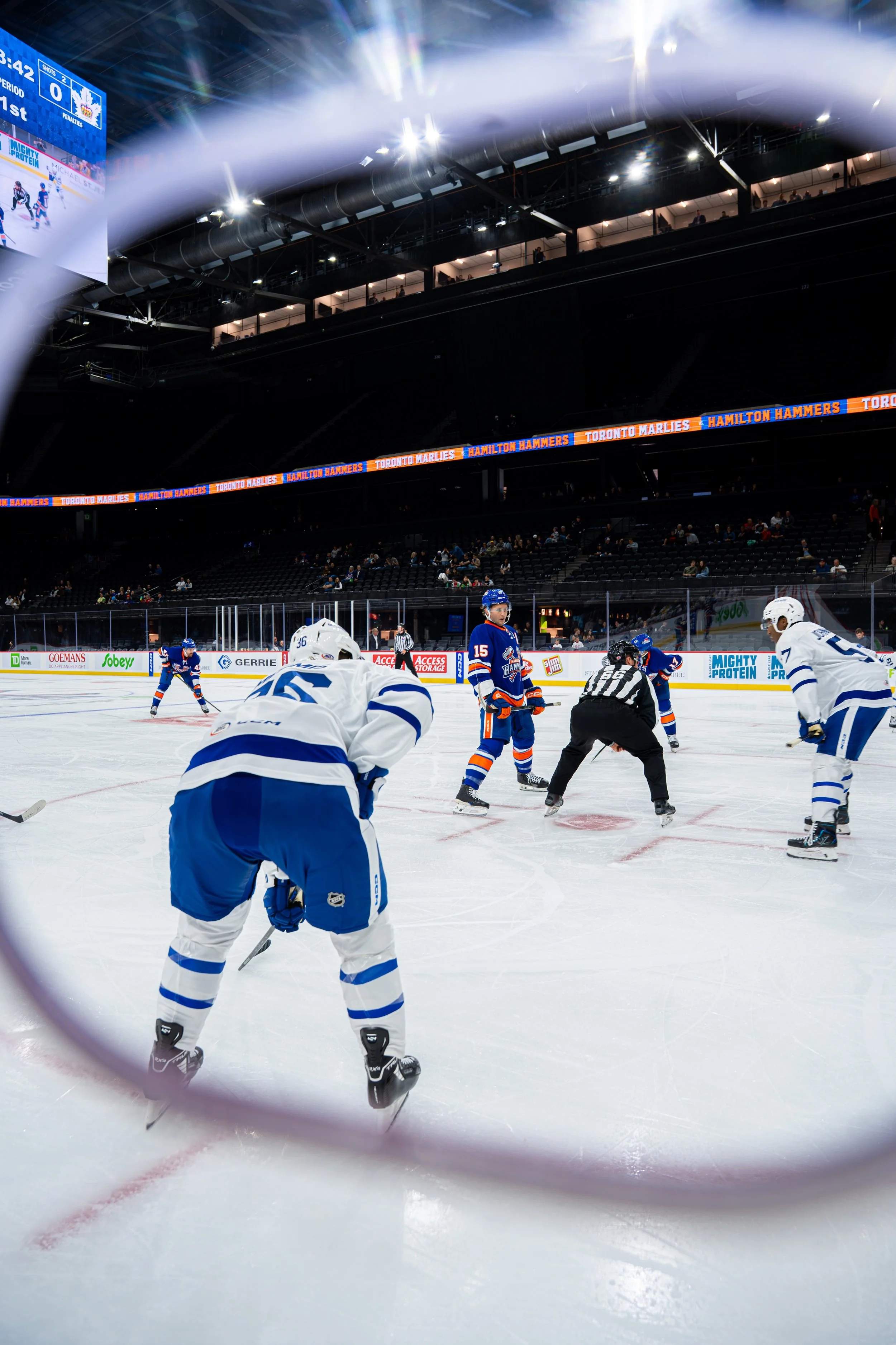 View through a hockey rink's protective glass shows players on the ice during a game, with a scoreboard and spectators in the stands in the background.