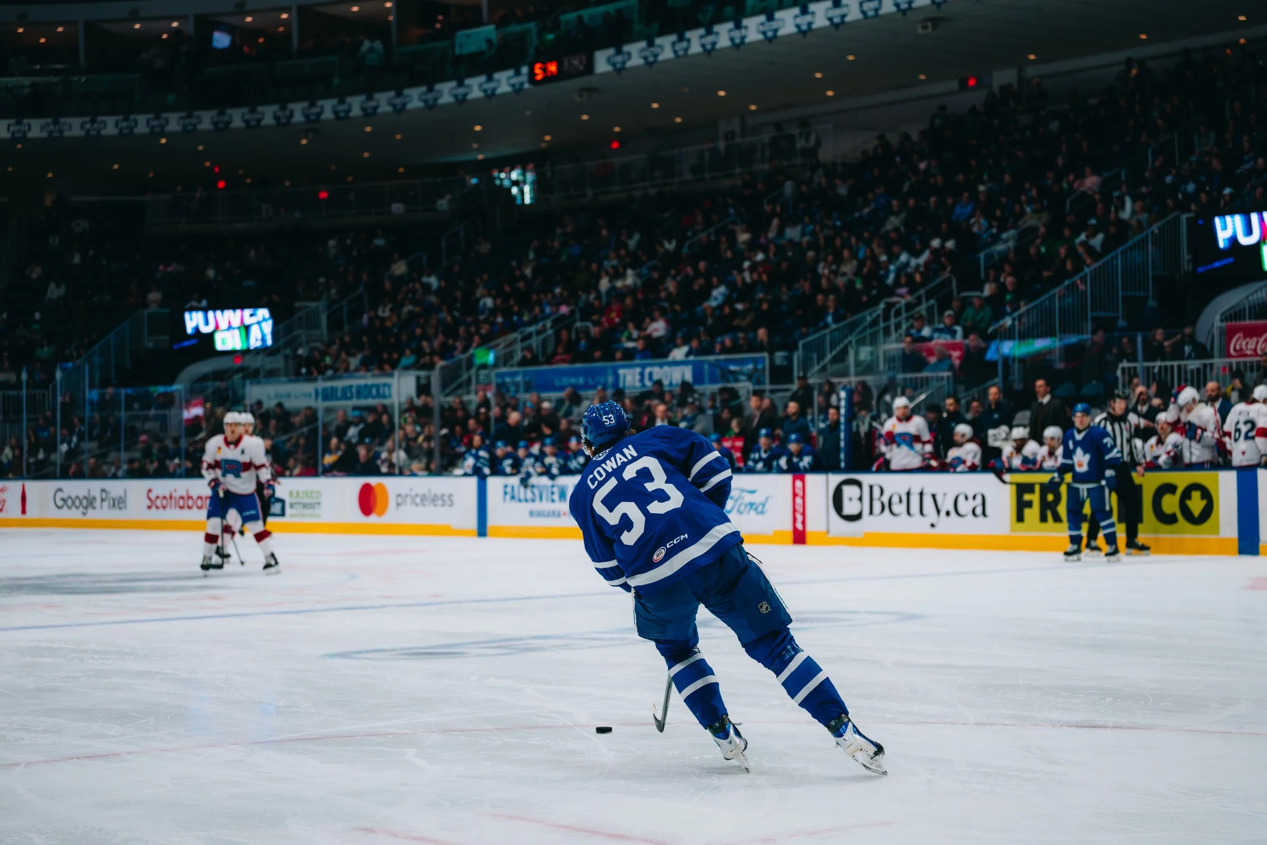 Ice hockey game with players on the ice rink and spectators in the stands, featuring a player in a blue jersey with the number 53 and the name Cowan.
