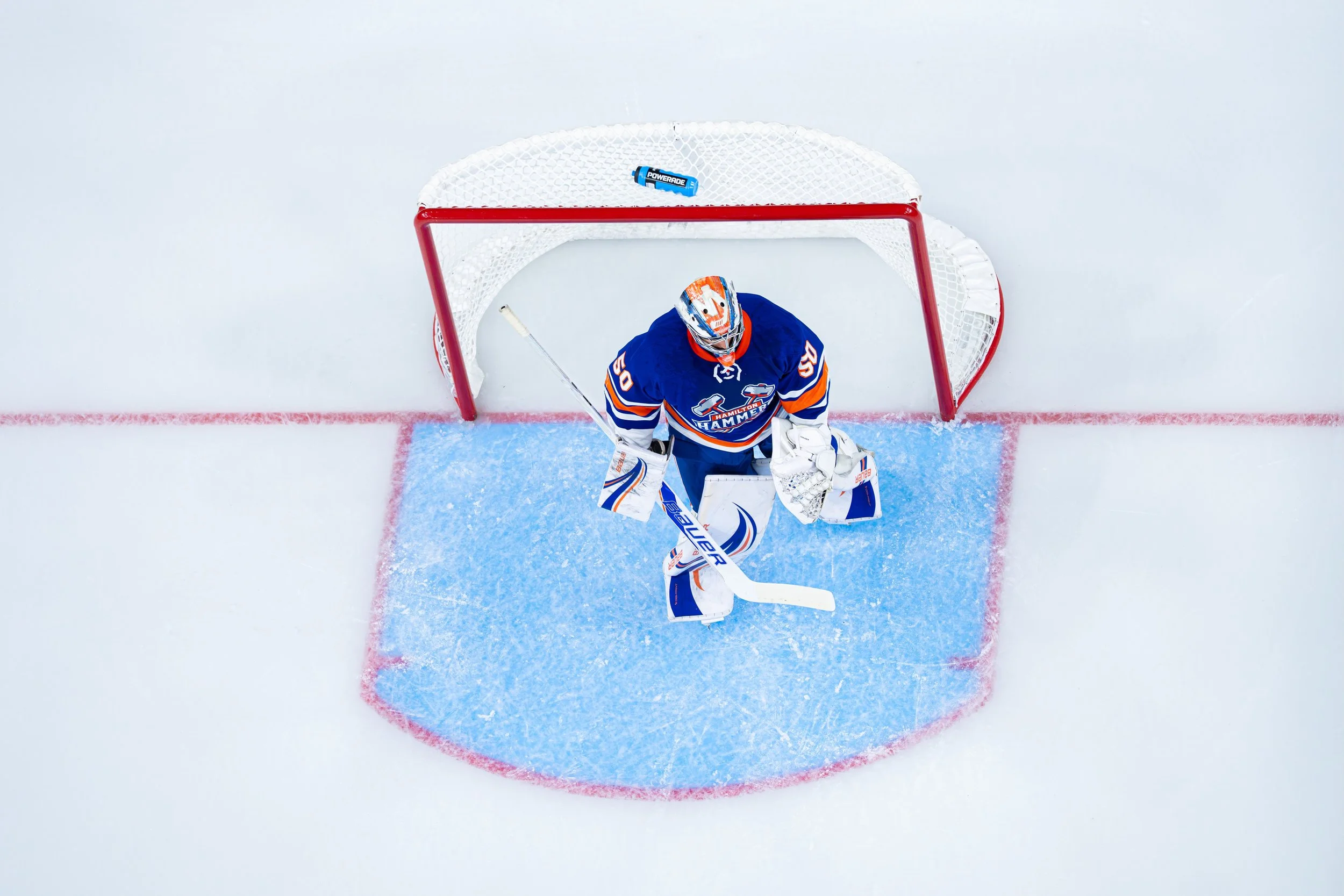 Overhead view of a hockey goalie standing in front of the goal on the ice, wearing blue and white gear with an orange face mask, holding a goalie stick, with a puck nearby.