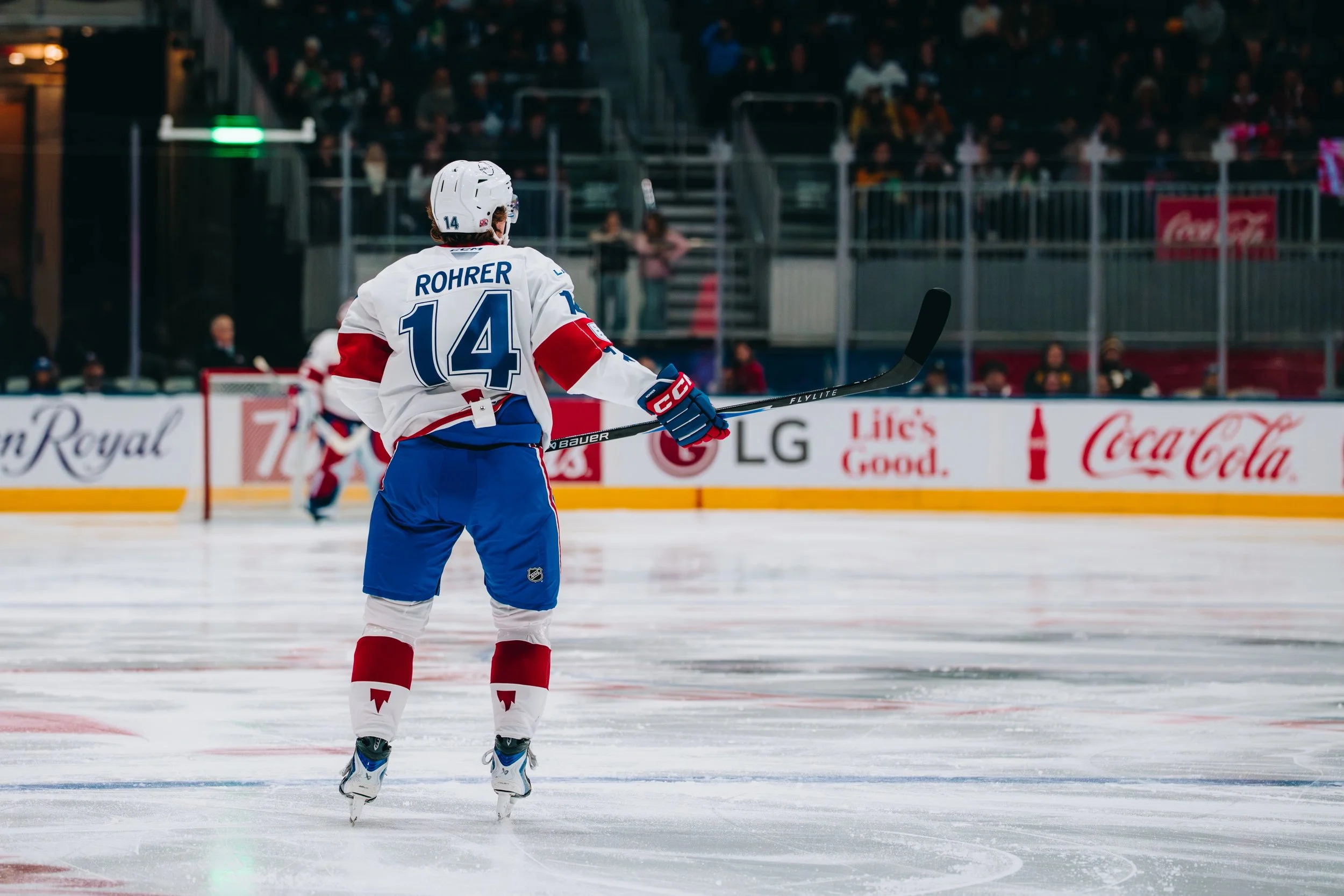 An ice hockey player wearing a white and red jersey with the name 'Rohrer' and number 14 on the back, standing on the ice rink with his stick in hand, facing away from the camera. The background shows spectators in the stands watching the game.