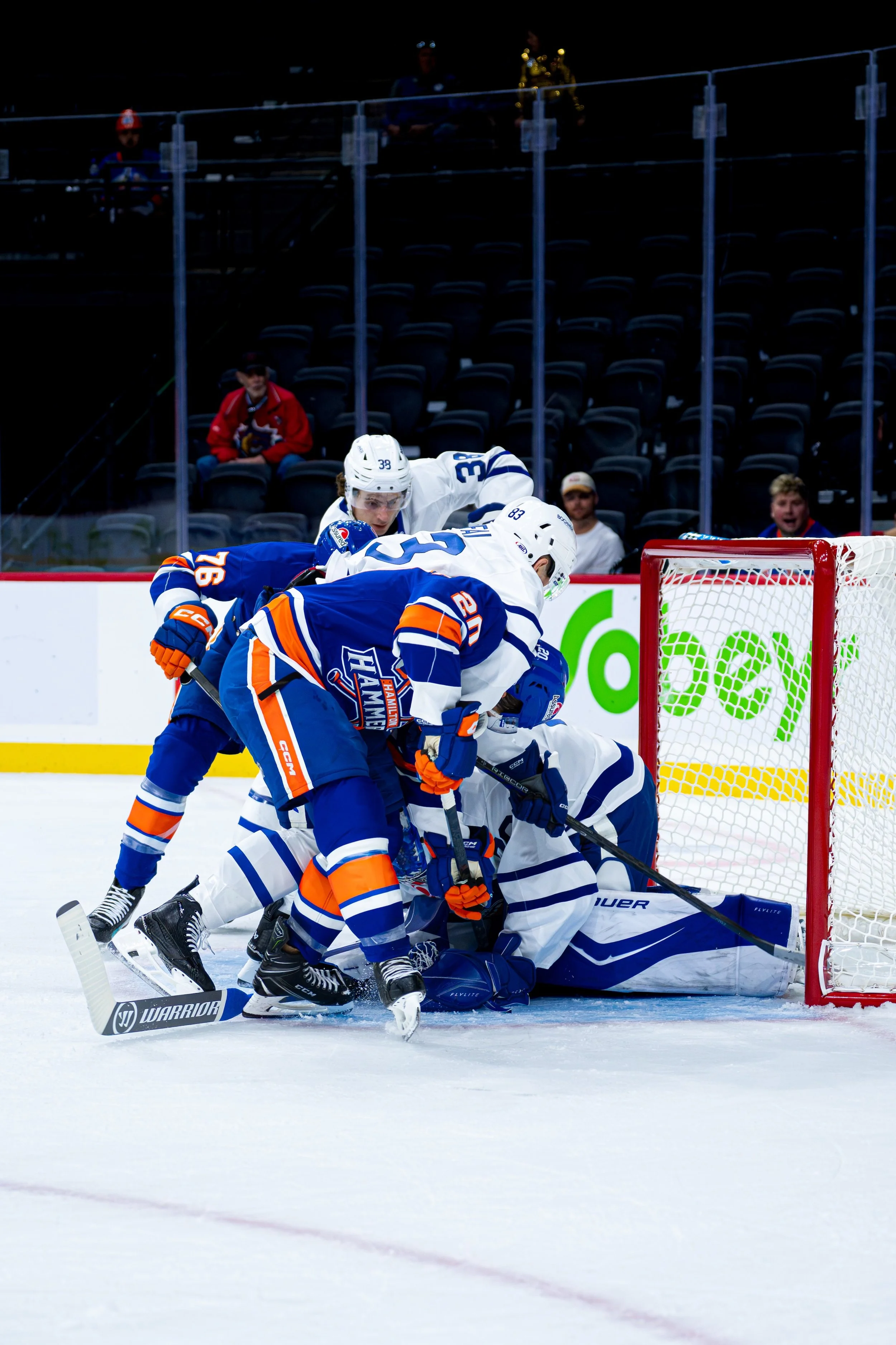 Hockey players are fighting for the puck near the goal in an ice hockey game, with empty seats and a few spectators watching in the background.