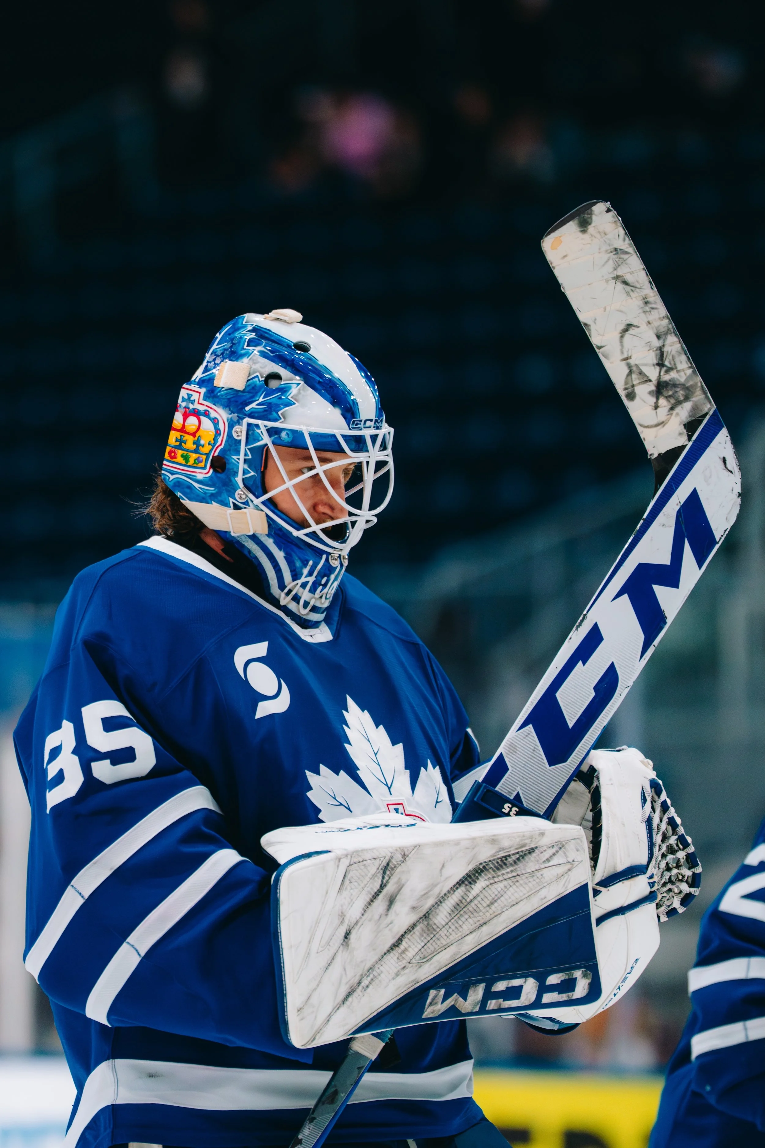 Ice hockey goalie wearing blue and white uniform and helmet, holding a hockey stick, standing on ice rink.