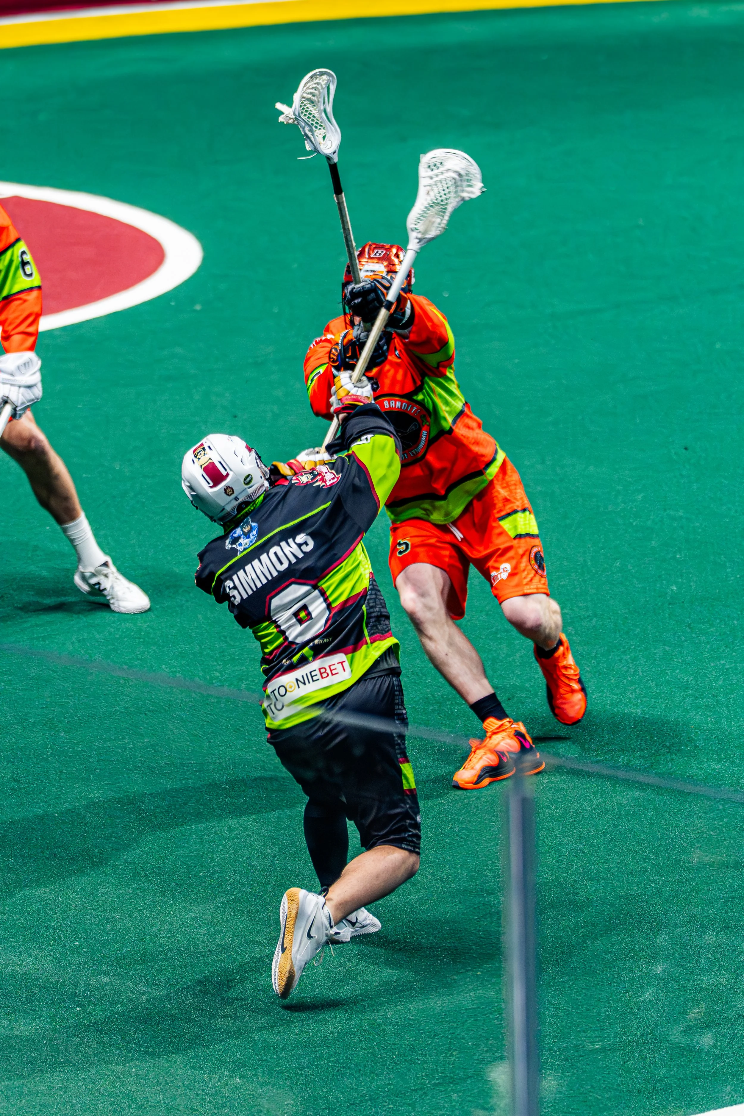 Two lacrosse players engaged in a game on a green turf field, with one player in an orange uniform defending and the other in a black uniform attempting to score or pass the ball.
