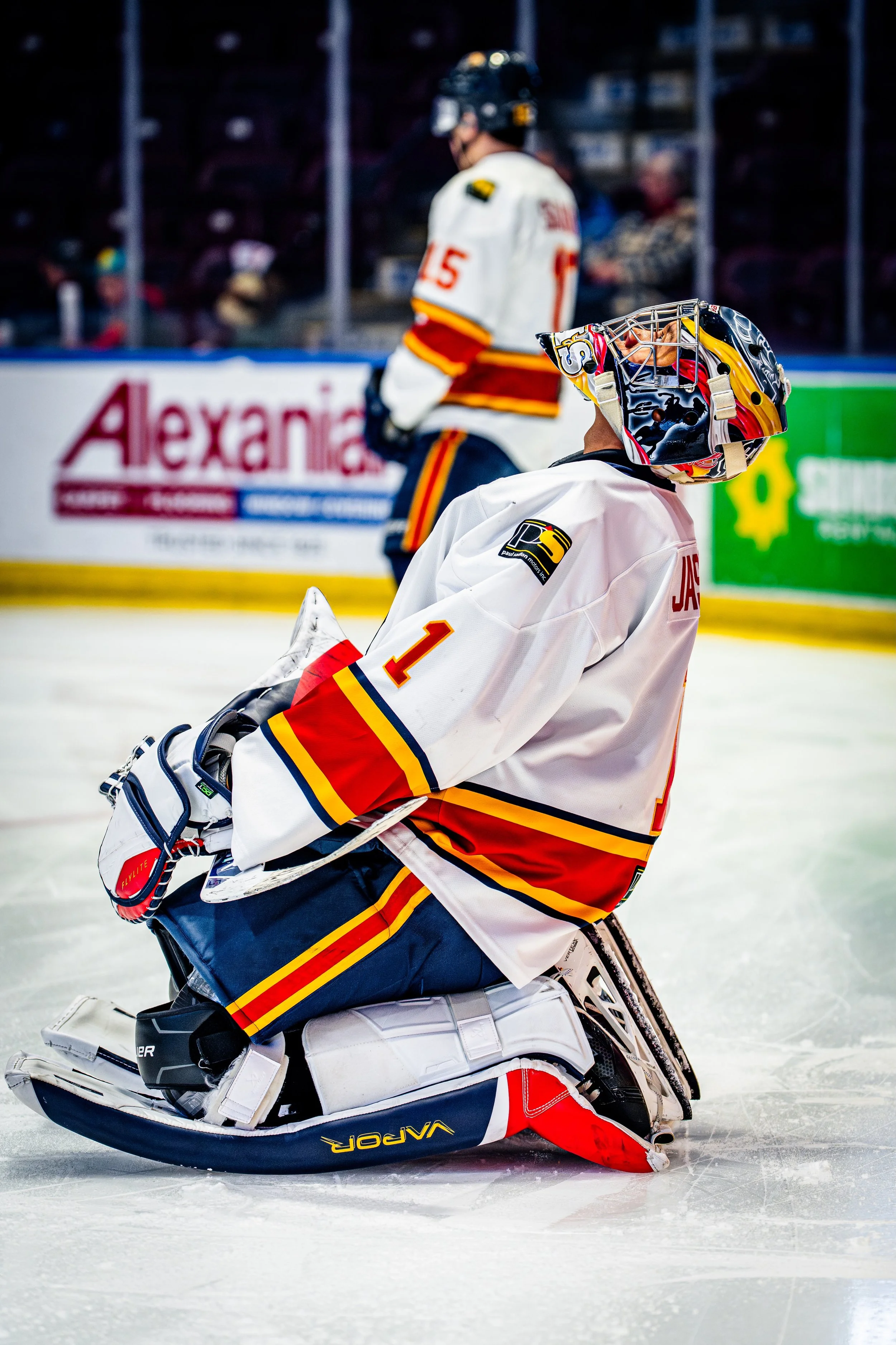 An ice hockey goalie in white, red, and yellow gear kneeling on the ice during a game, with another player in the background and spectators in the stands.