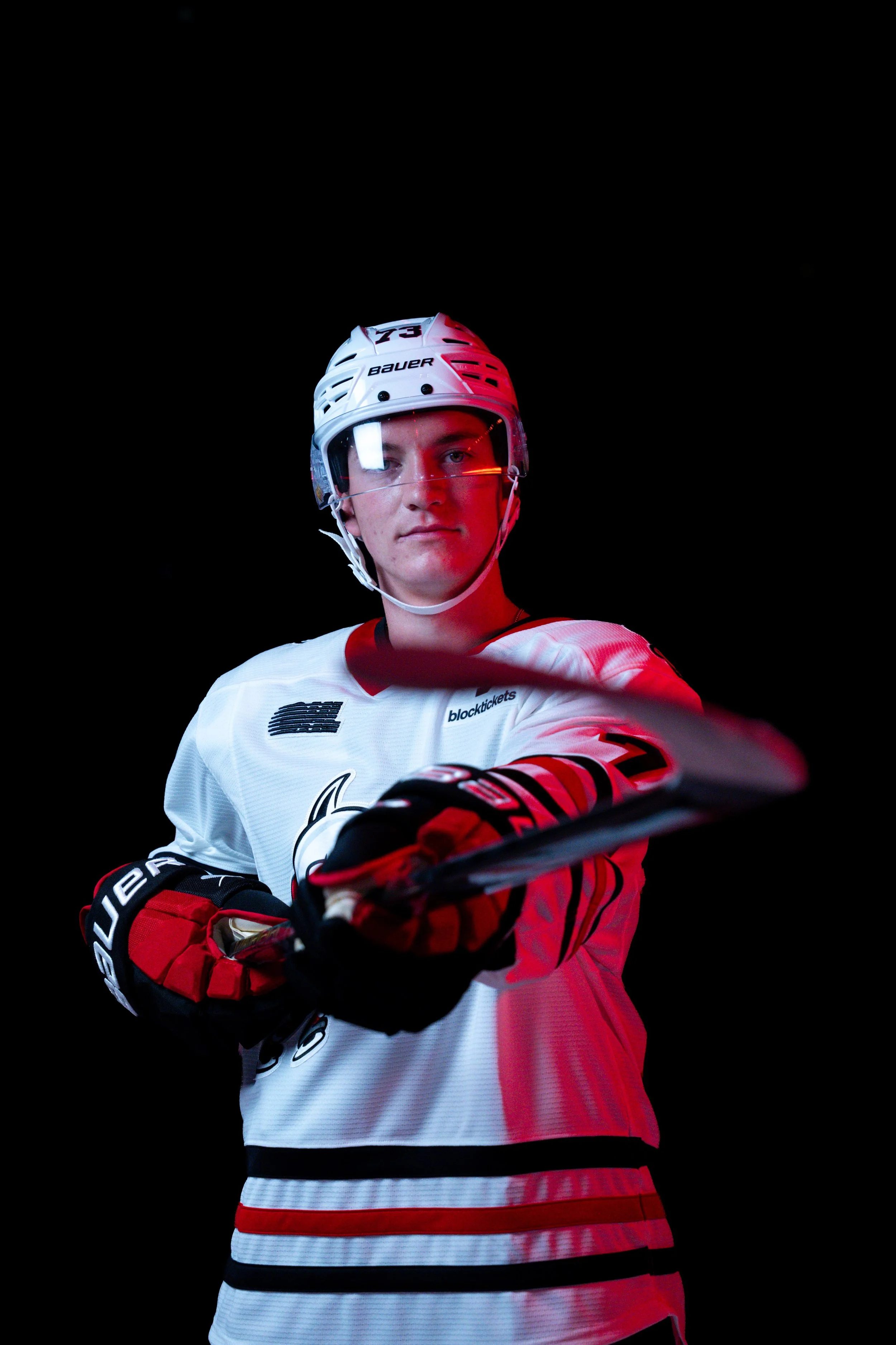 A young male hockey player wearing a white and red jersey with black and red gloves, a white helmet with a clear visor, and holding a hockey stick, posing against a black background.