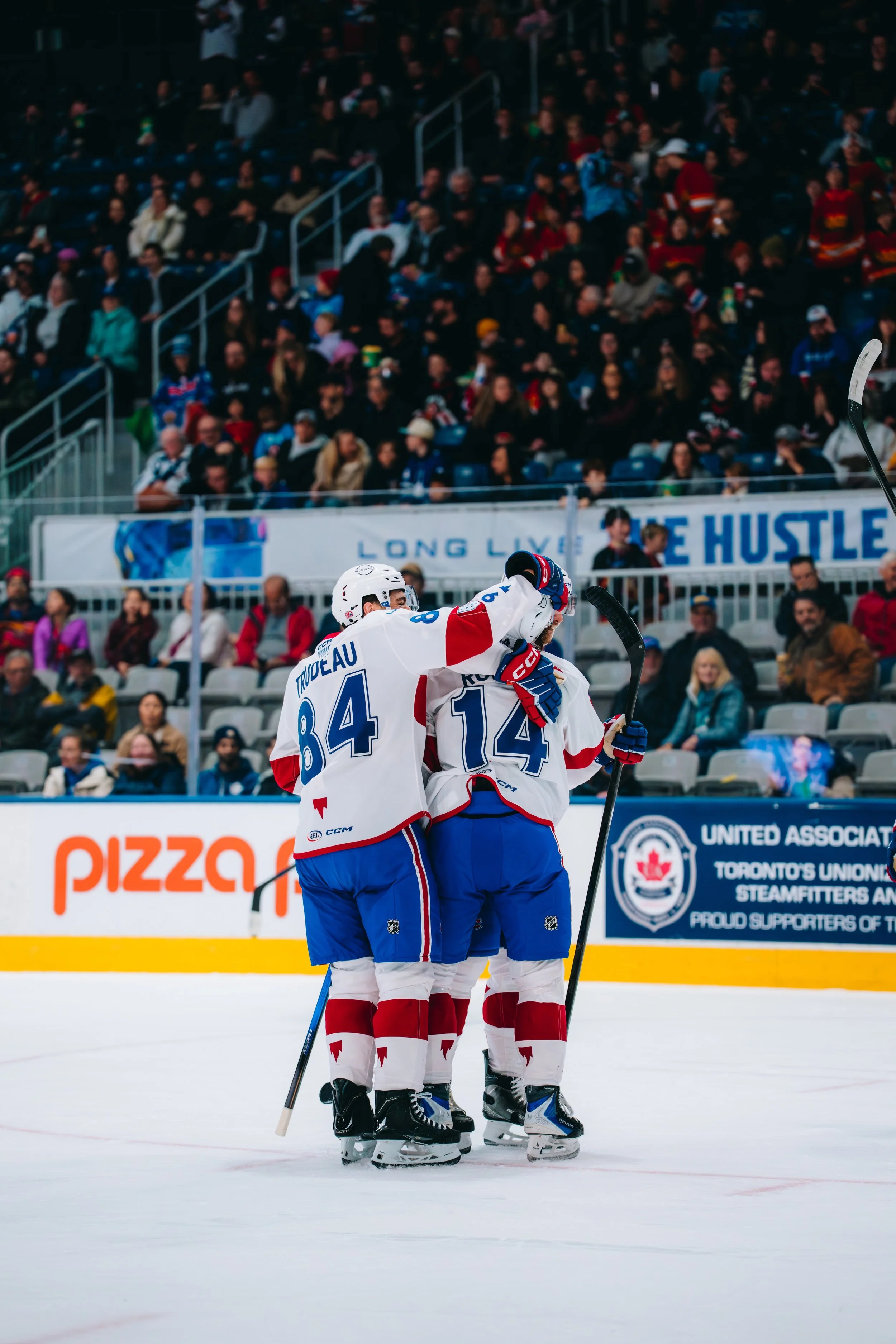 Two ice hockey players in red, white, and blue uniforms hugging on the ice rink. In the background, a crowded arena crowd observes the celebration.