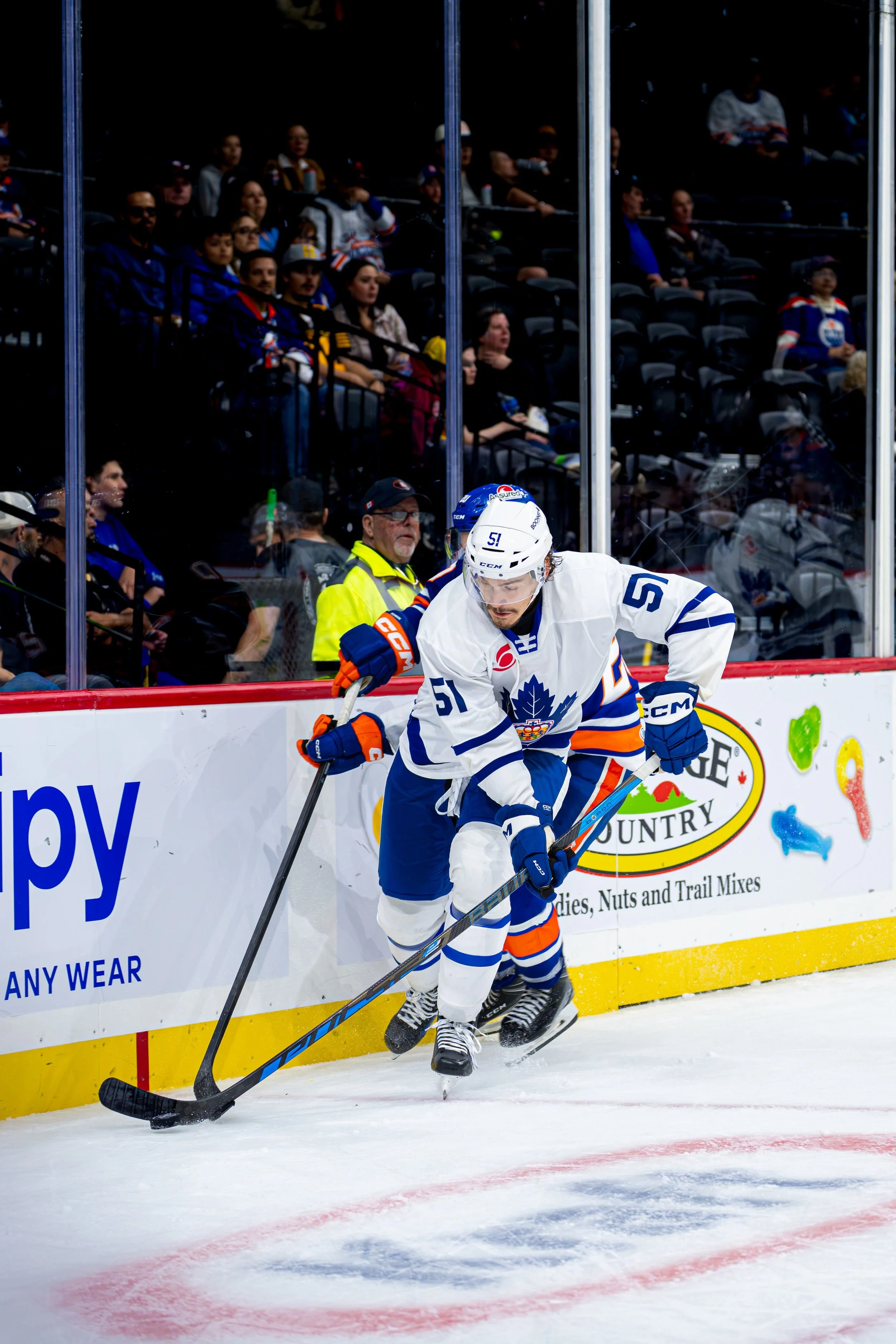 Hockey player in a white jersey with a blue maple leaf logo and number 51, skating along the rink wall, with fans watching from behind the glass.