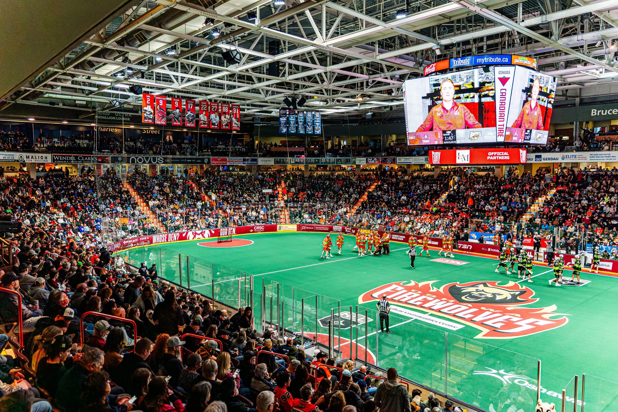 An indoor sports arena filled with spectators watching a hockey game. Players are gathered on the rink with a large digital scoreboard above showing a close game. Red and blue banners hang from the ceiling.