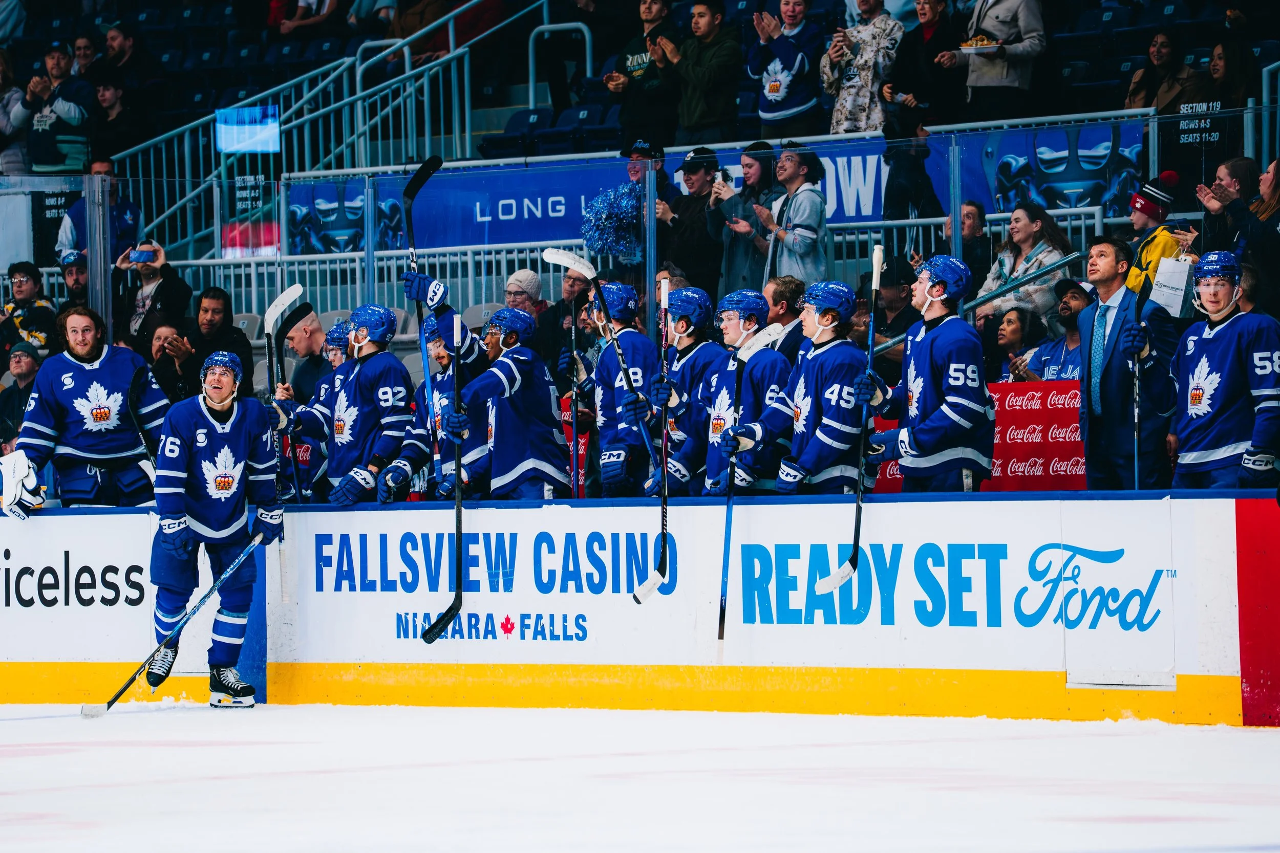An NHL hockey team in blue jerseys standing on the ice rink, with spectators in the stands clapping and cheering behind a glass barrier. Some fans are taking photos, and others are watching the game. Advertisement banners and arena signage are visibl
