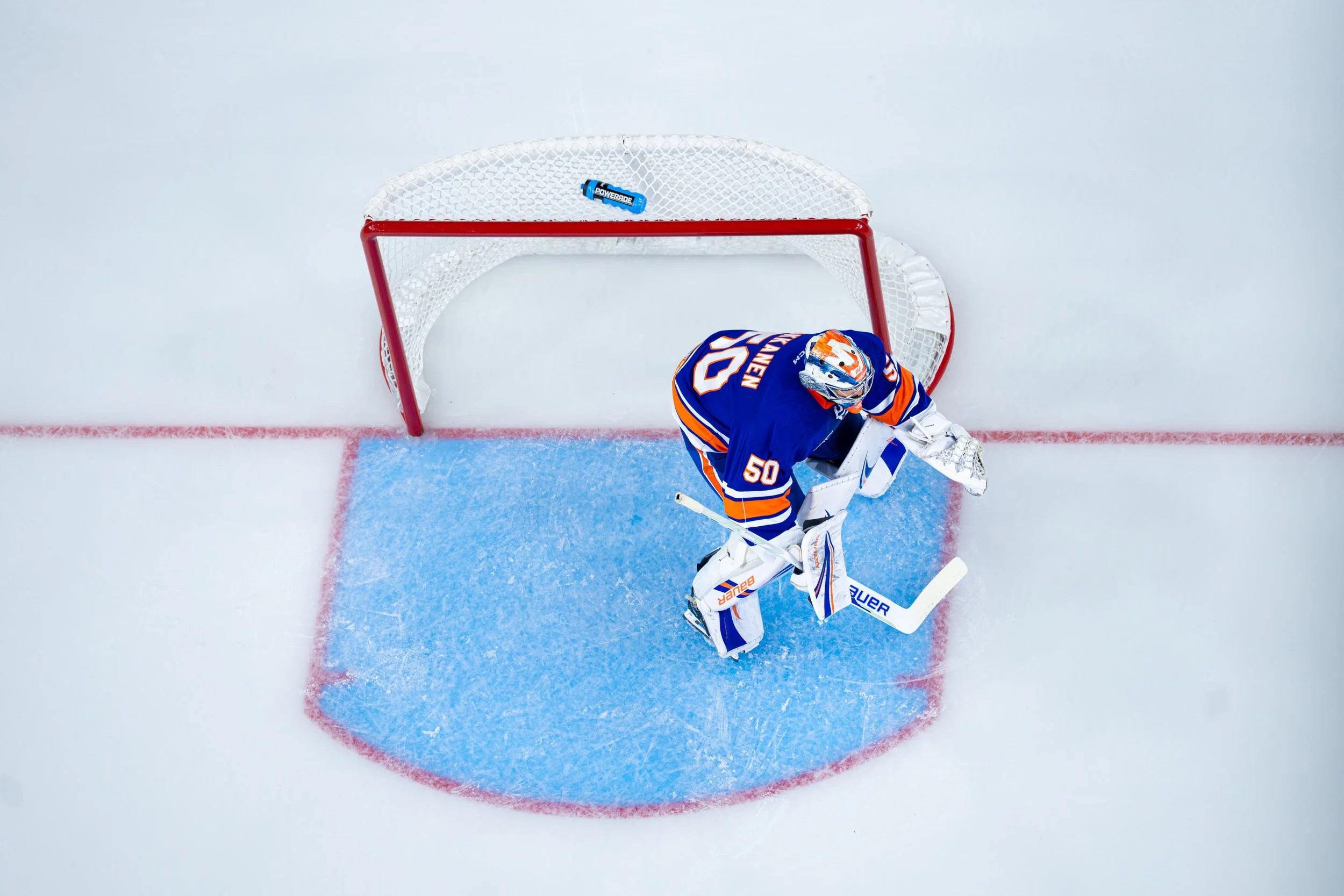 Overhead view of an ice hockey goalie in blue and orange uniform making a save in front of the net, with a Powerade bottle on top of the net.