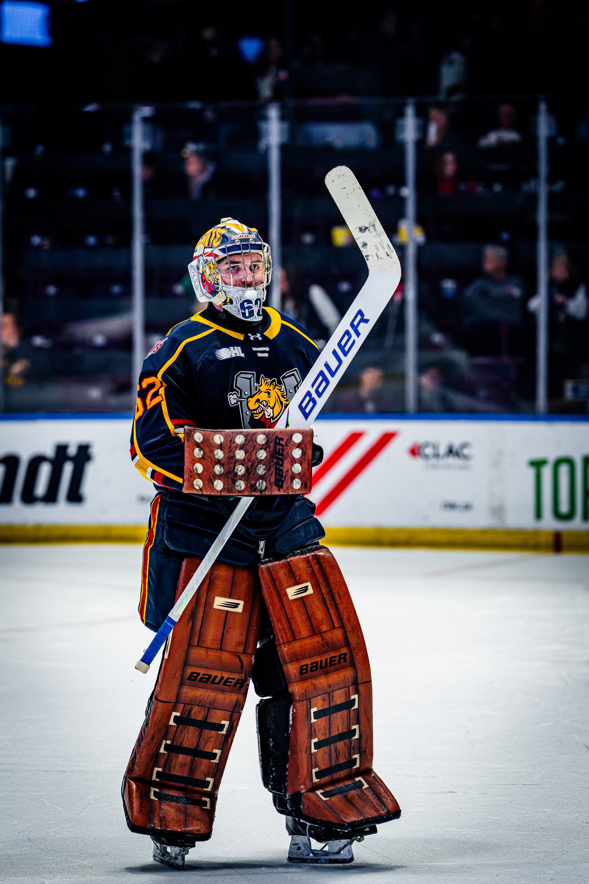 A hockey goaltender standing on the rink, wearing a helmet, jersey, and protective gear, holding a goalie stick. In the background, spectators are watching the game from behind the glass.