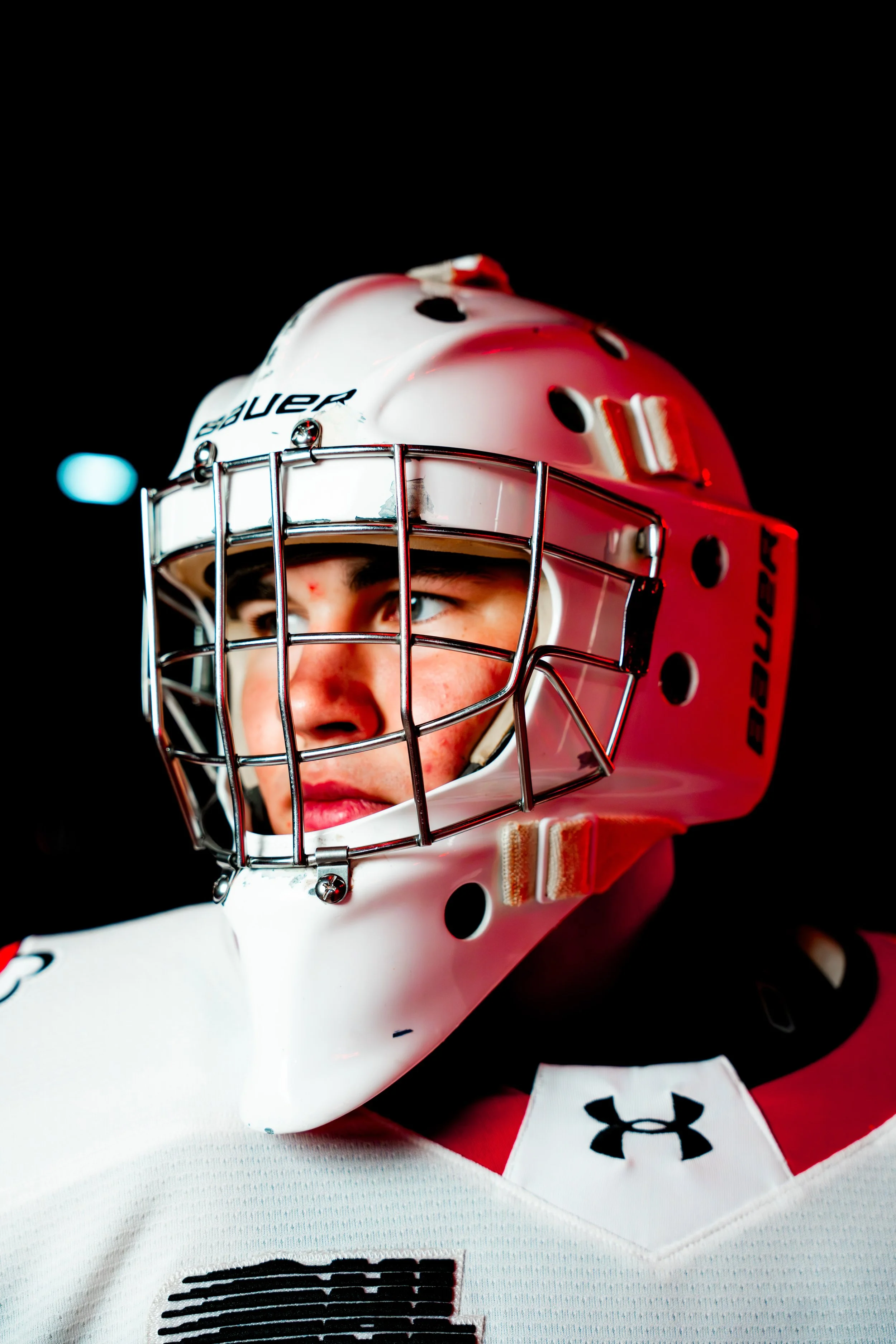 Close-up of an ice hockey goalie in full gear, wearing a white helmet with a red chin guard and a metal cage, and a white jersey with black and red accents.