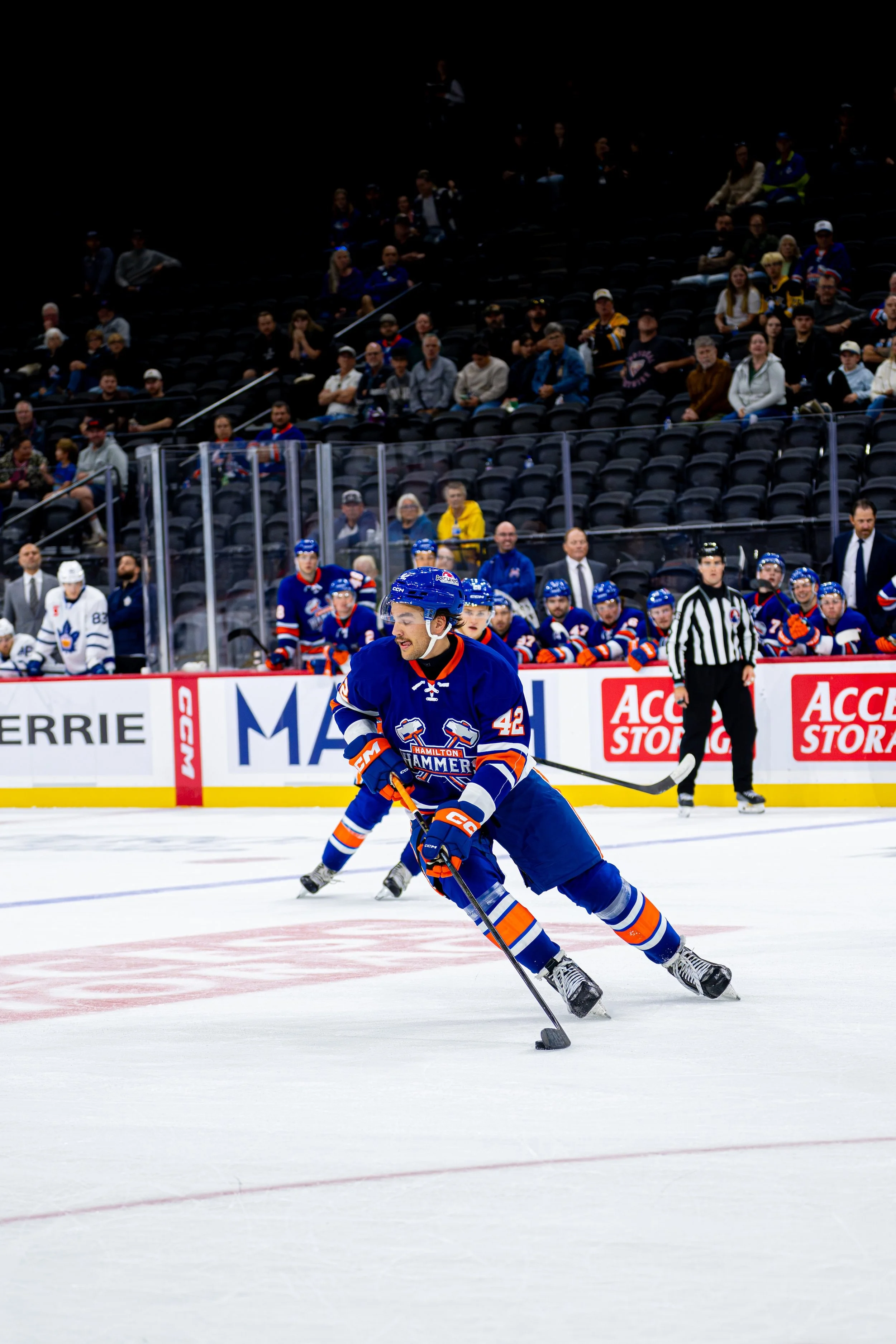 Ice hockey game with players in blue and white jerseys on the ice rink, spectators seated in the background.