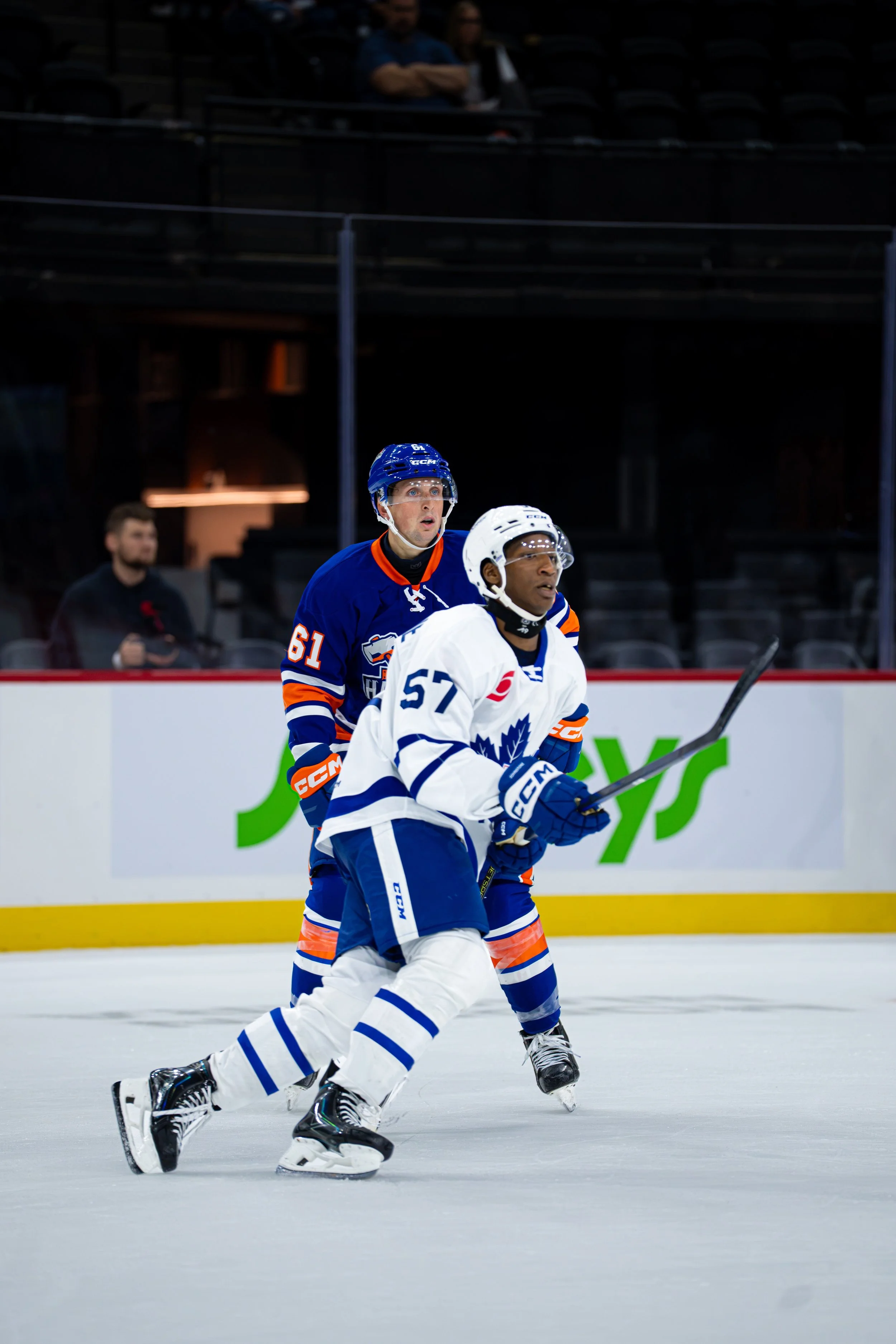 Two ice hockey players skating on the ice during a game, with one in a white jersey and the other in a blue jersey, and spectators in the background.