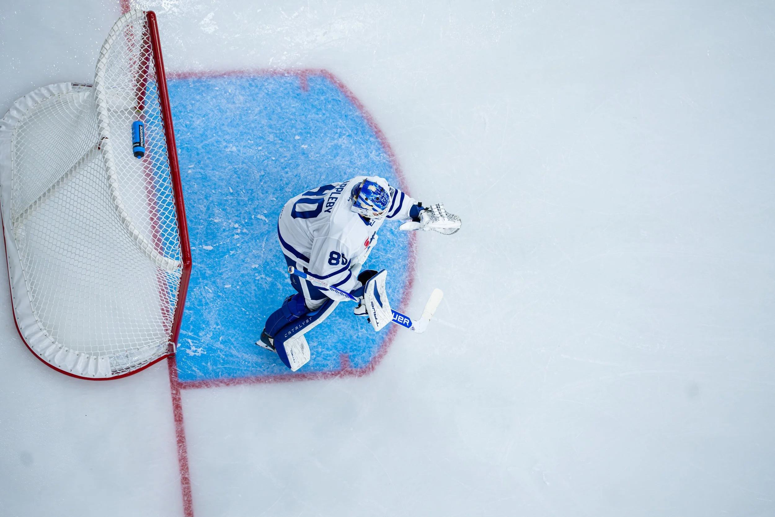Hockey goalie in white and blue gear on ice, positioned near goal with net, in the midst of a game.