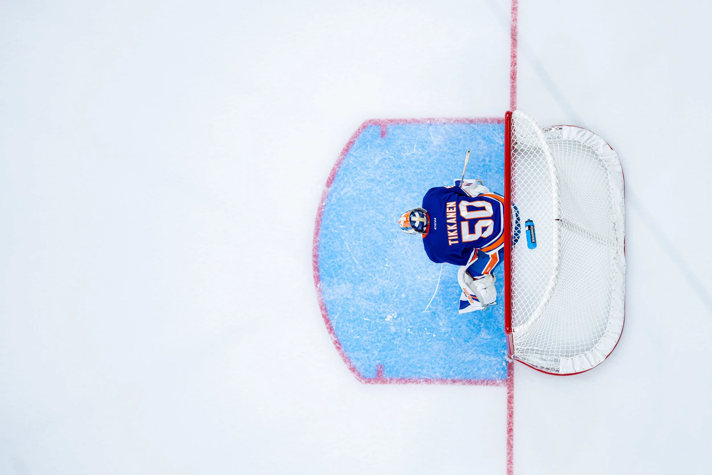 An ice hockey goalie in a blue jersey and helmet sitting on the ice in front of their net, with equipment and a hockey stick nearby.