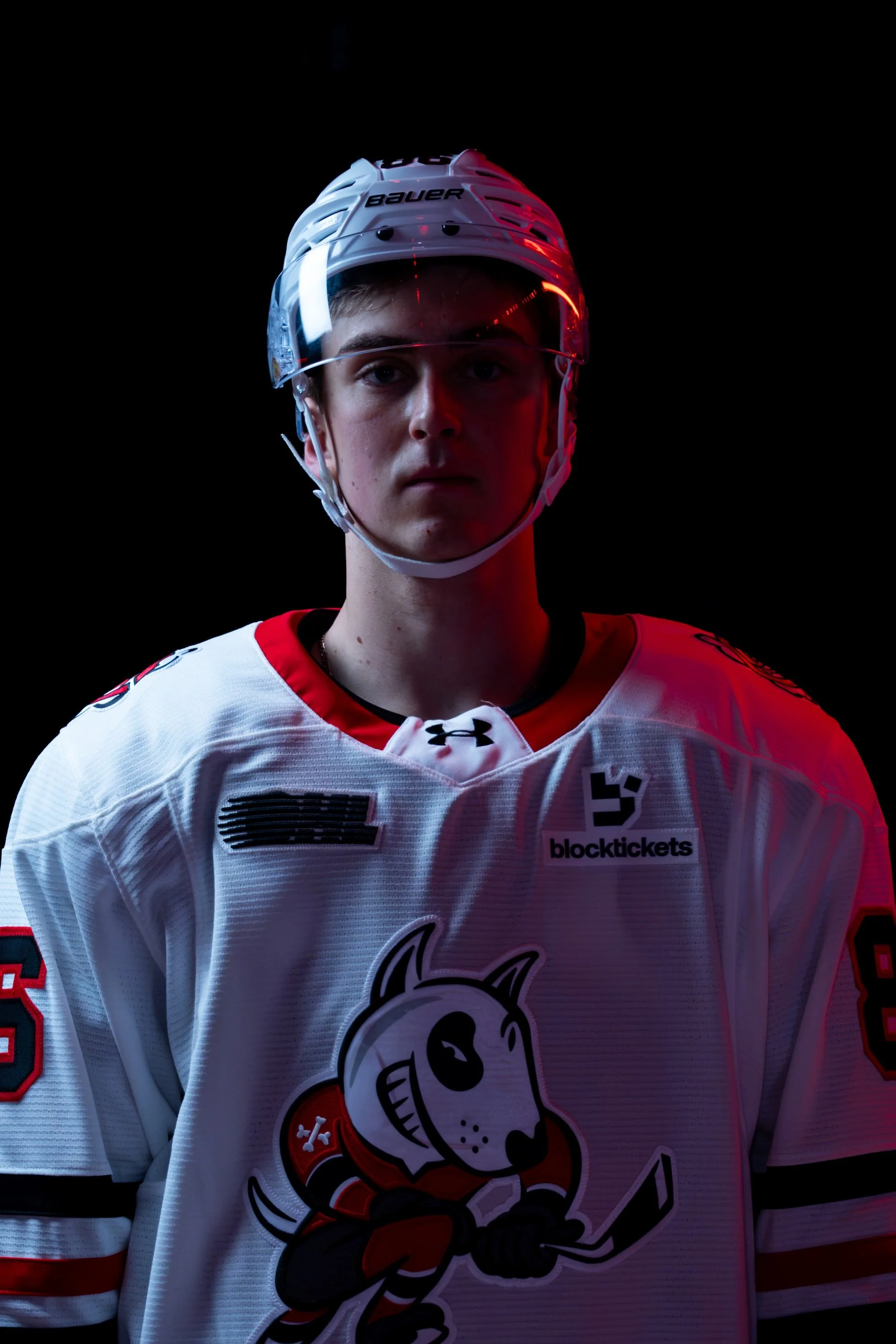 Close-up of a young hockey player wearing a white jersey with a cartoon mascot and various sponsor logos, including 'blocktickets'. The player is wearing a white helmet with a clear visor, and has short hair. The background is black.