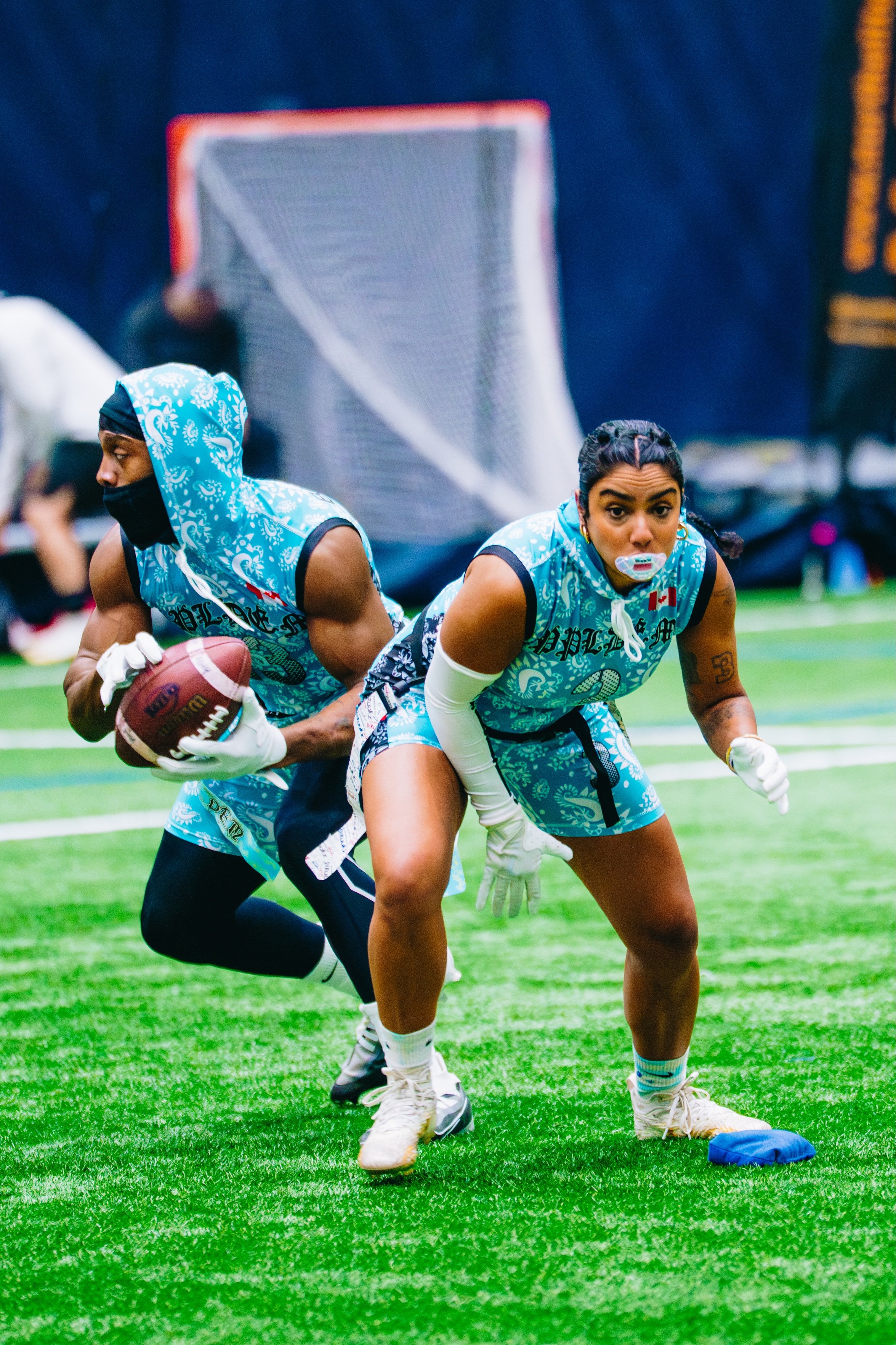 Two women in blue athletic outfits with black accents participating in a football drill on a green turf field, one holding a football and the other in a ready stance, with football gear and a goal net in the background.