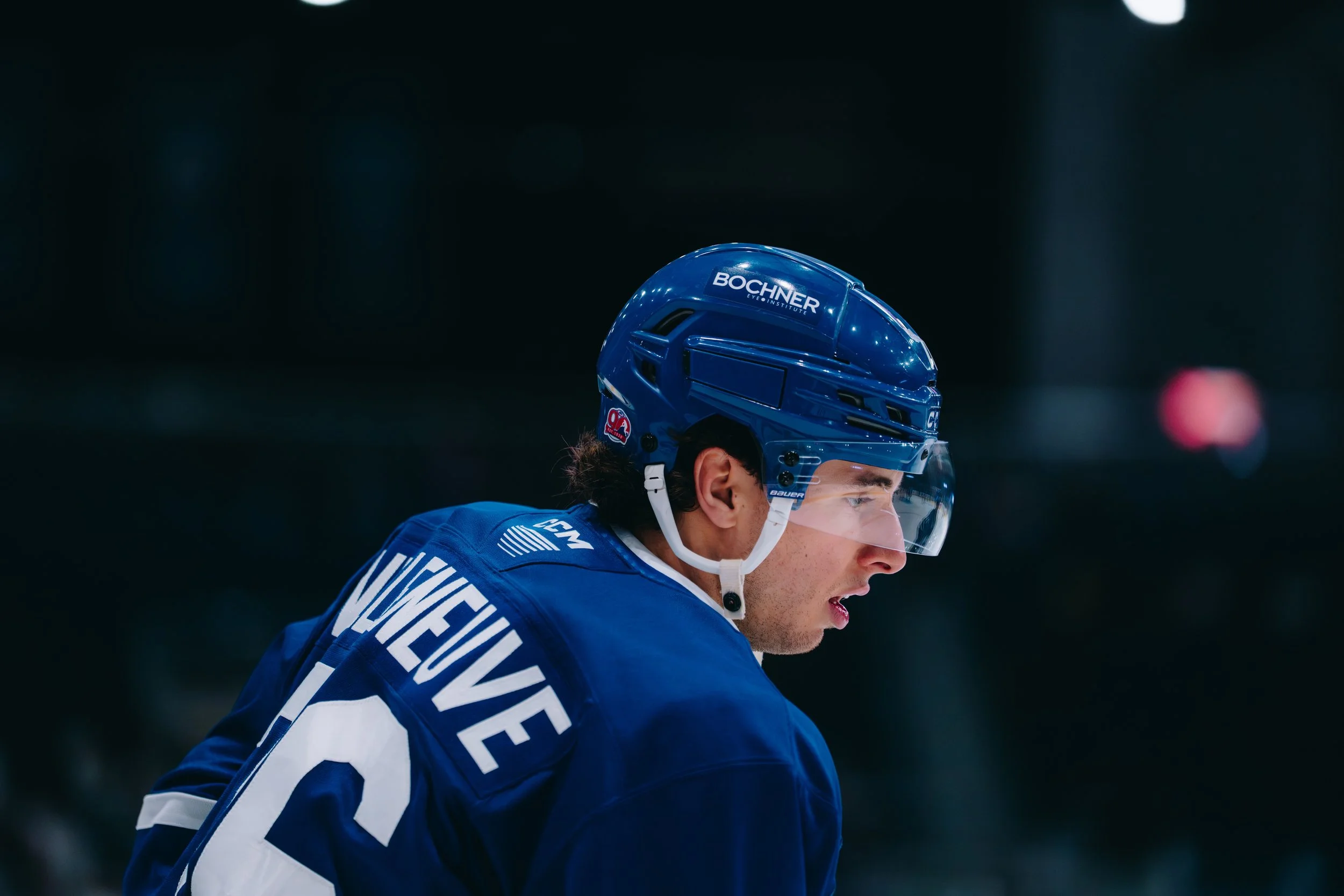 Close-up of a hockey player wearing a blue helmet with "BCHNER" logo and a blue jersey, in an indoor ice rink.