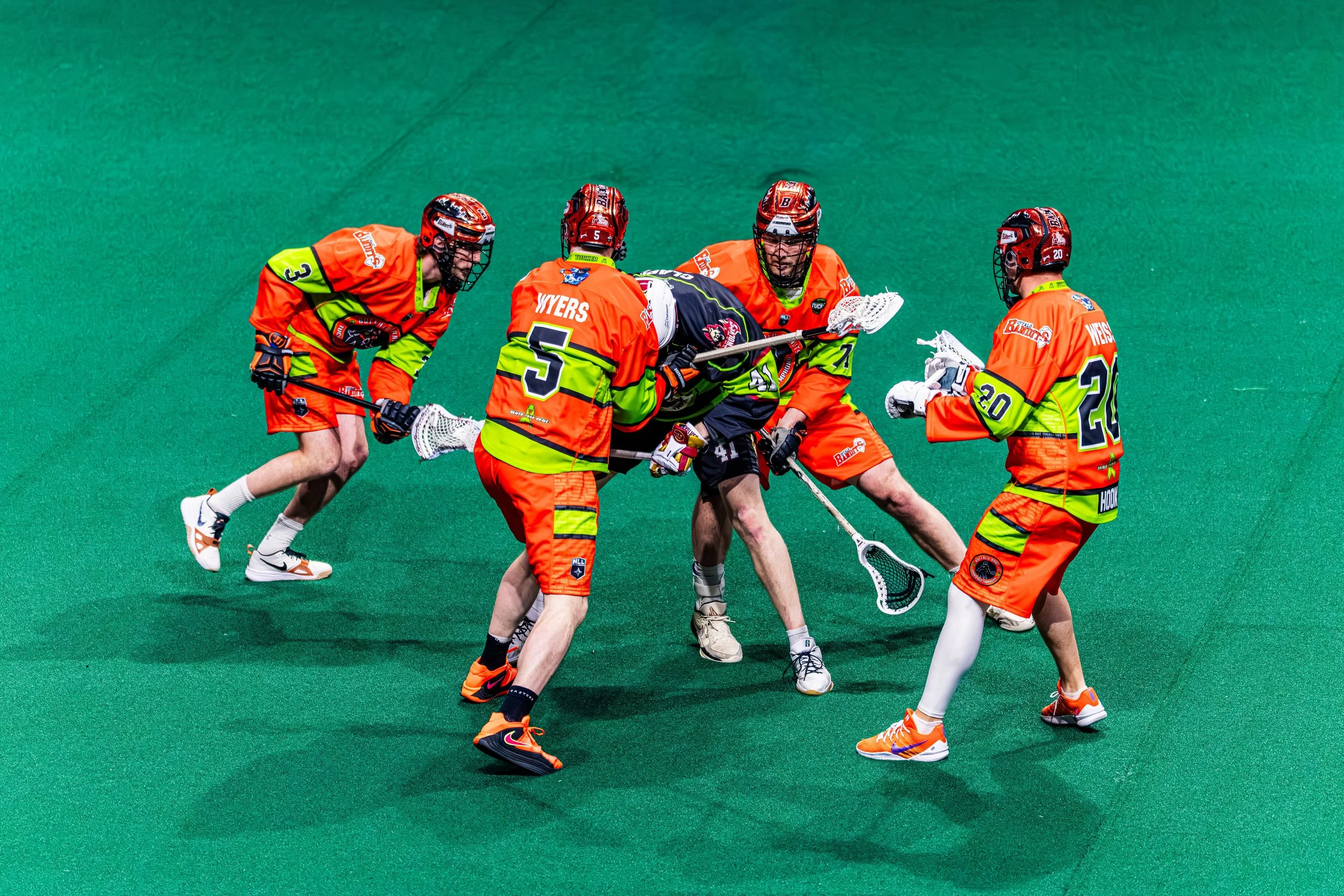 A group of male field hockey players in bright orange uniforms on a green turf, engaged in a play or celebration.