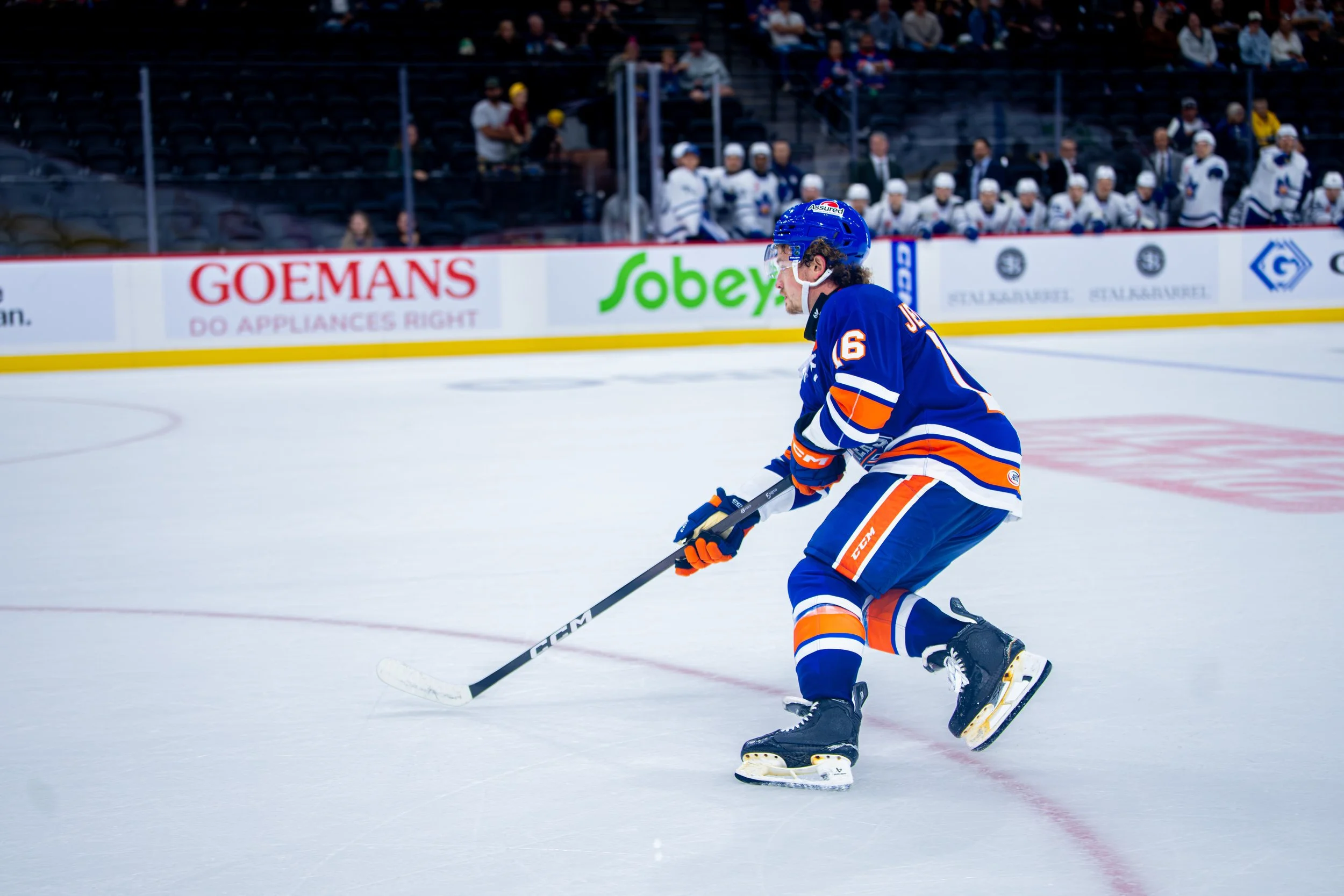 Ice hockey player in blue jersey skating on the ice during a game, with spectators in the background.