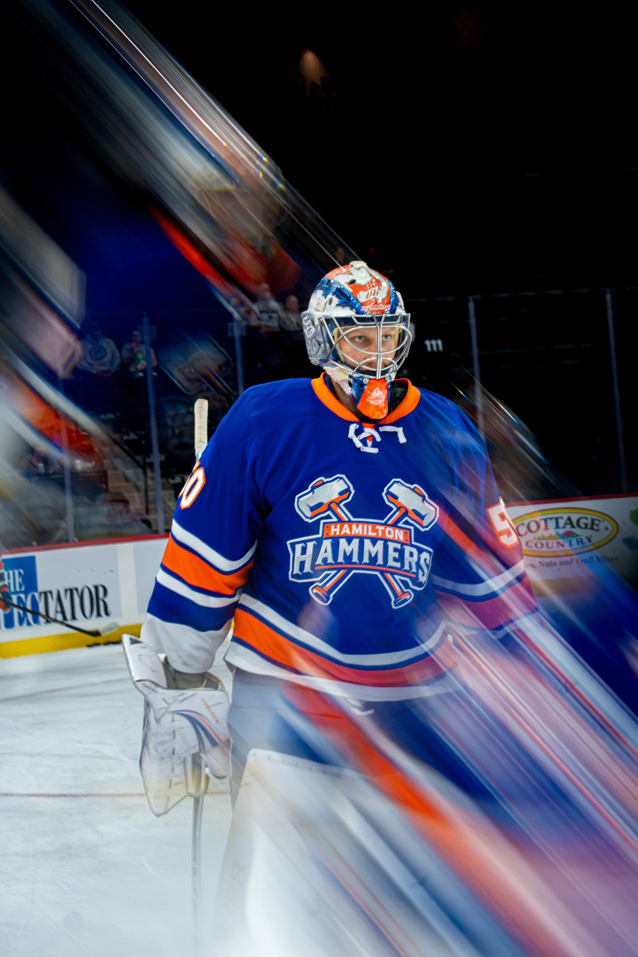 An ice hockey goalie wearing a blue and orange jersey with the logo 'Hamilton Hammers' stands on the ice rink looking at the camera. The goalie is wearing a helmet and gloves, with a dark background and blurred audience and advertisements behind.