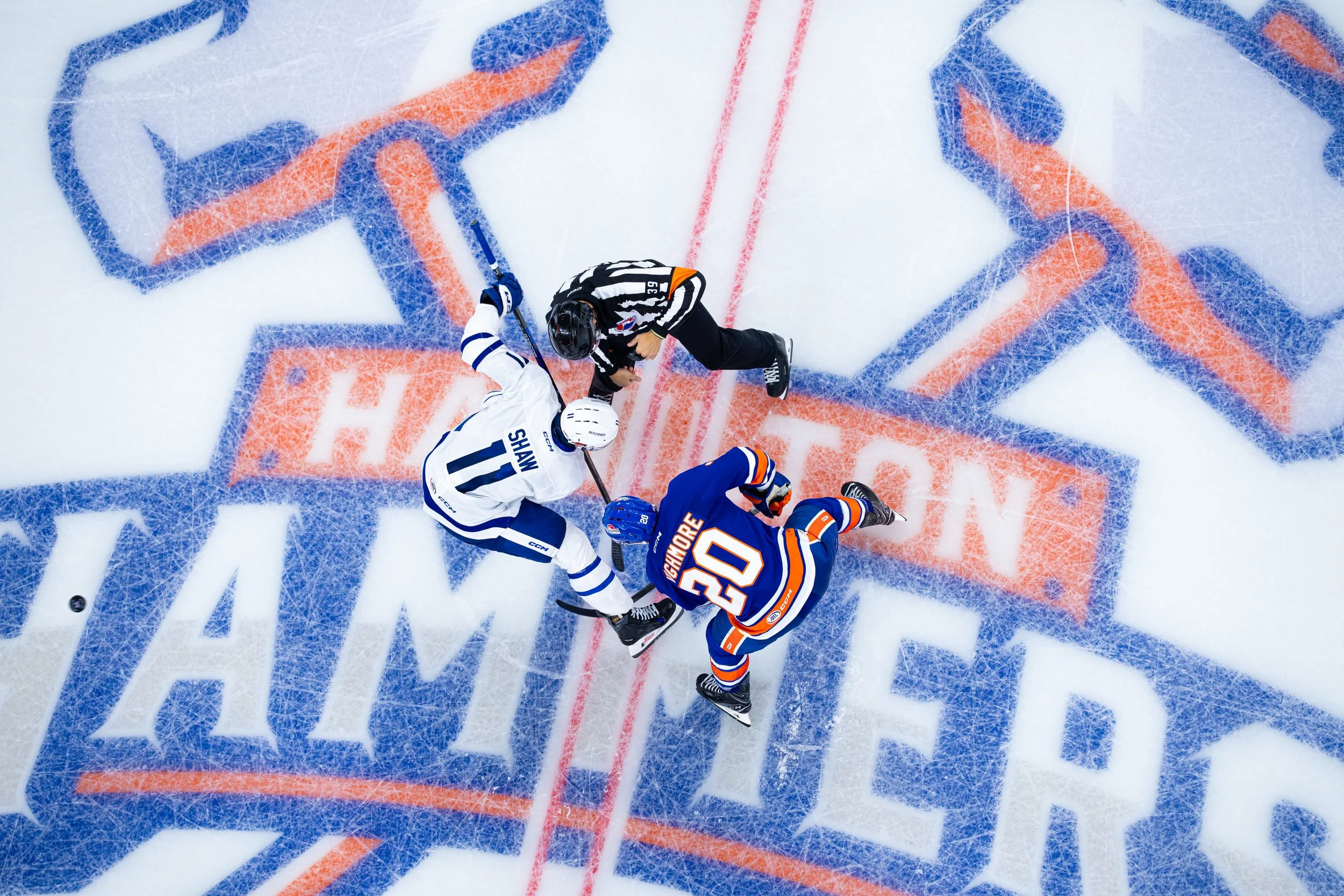 An overhead view of a hockey game face-off at center ice with players from two teams, one in white and the other in orange and blue, and a referee preparing to start the game.