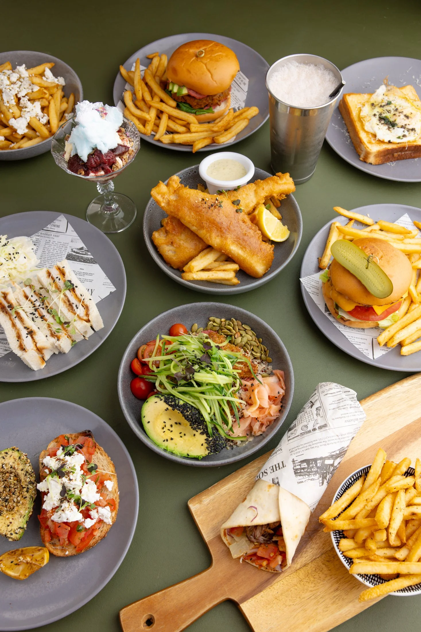 Assorted plates of food including burgers, fries, fish and chips, salad, flatbread, and dessert on a green table at a restaurant.