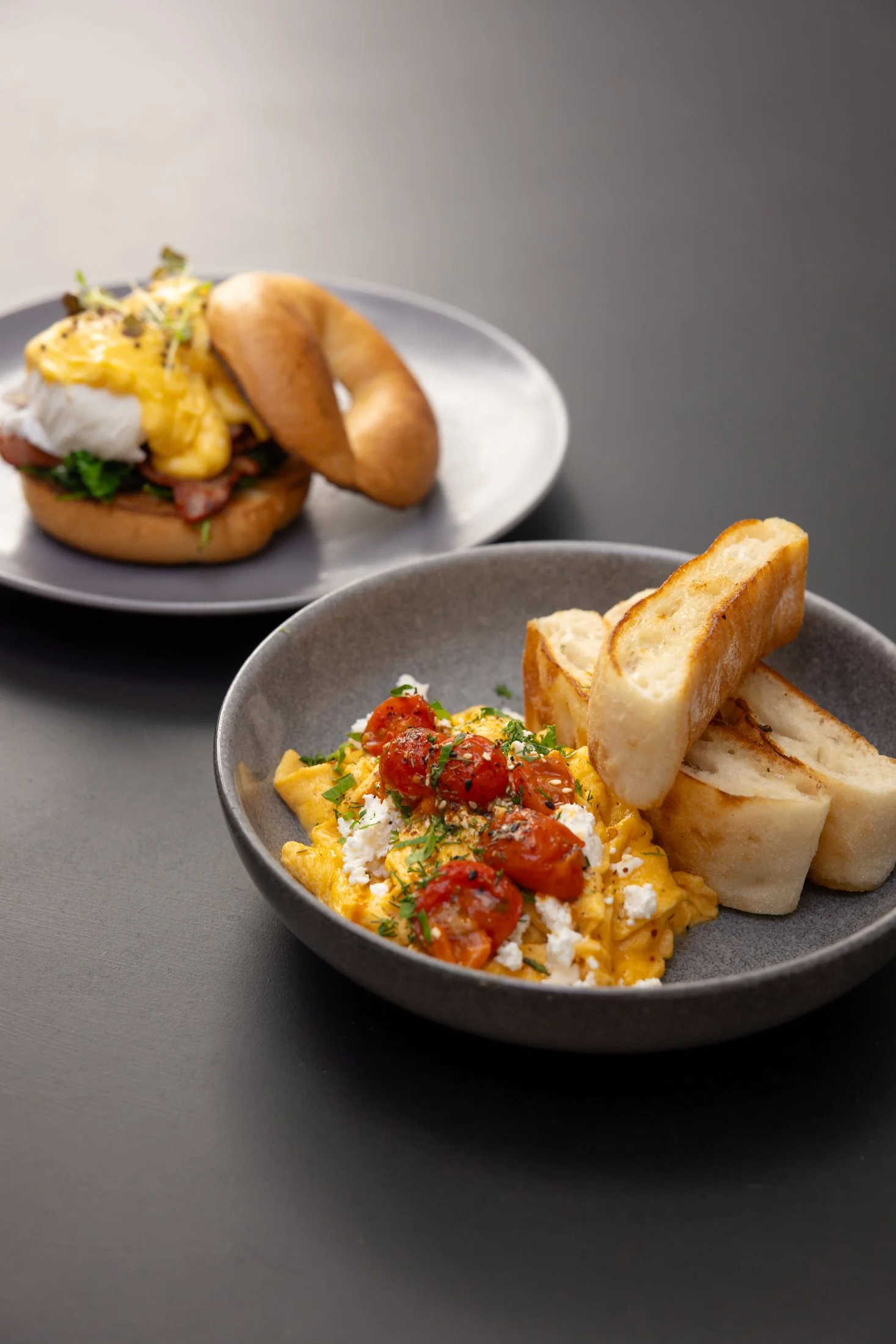 A plate of scrambled eggs with cherry tomatoes, crumbled cheese, herbs, and toasted bread slices, with a breakfast sandwich on a separate plate in the background.