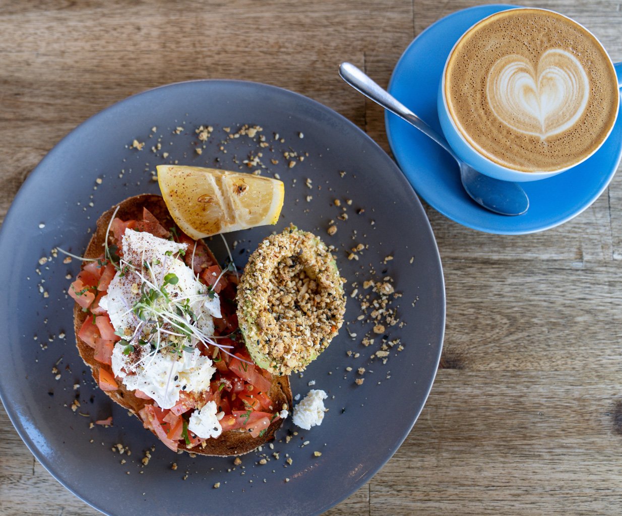 Plate with avocado, poached egg, tomato on toast, lemon wedge, and latte on a blue saucer.