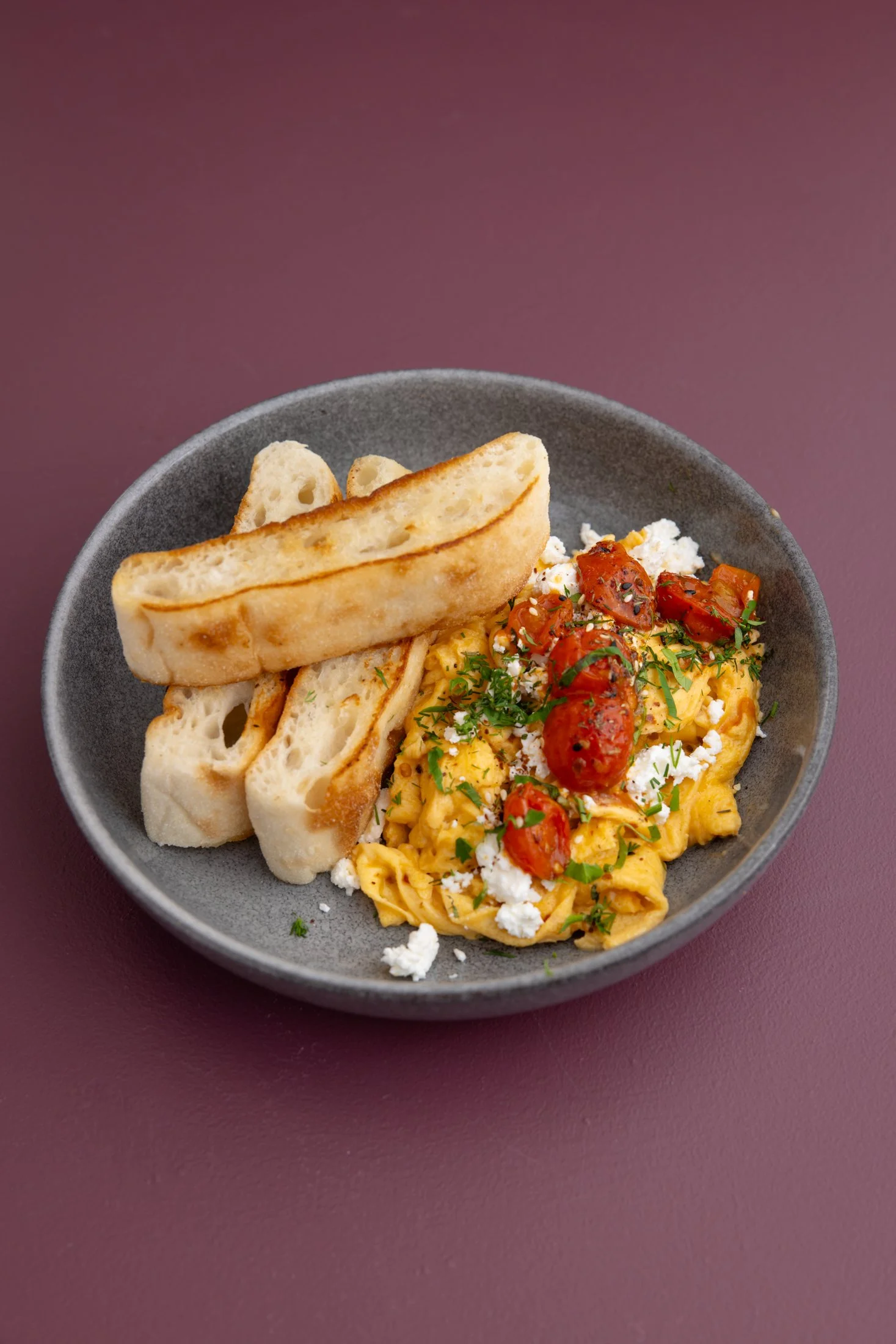 A gray bowl holding scrambled eggs topped with cherry tomatoes, herbs, and cheese, served with toasted slices of bread, all on a maroon surface.