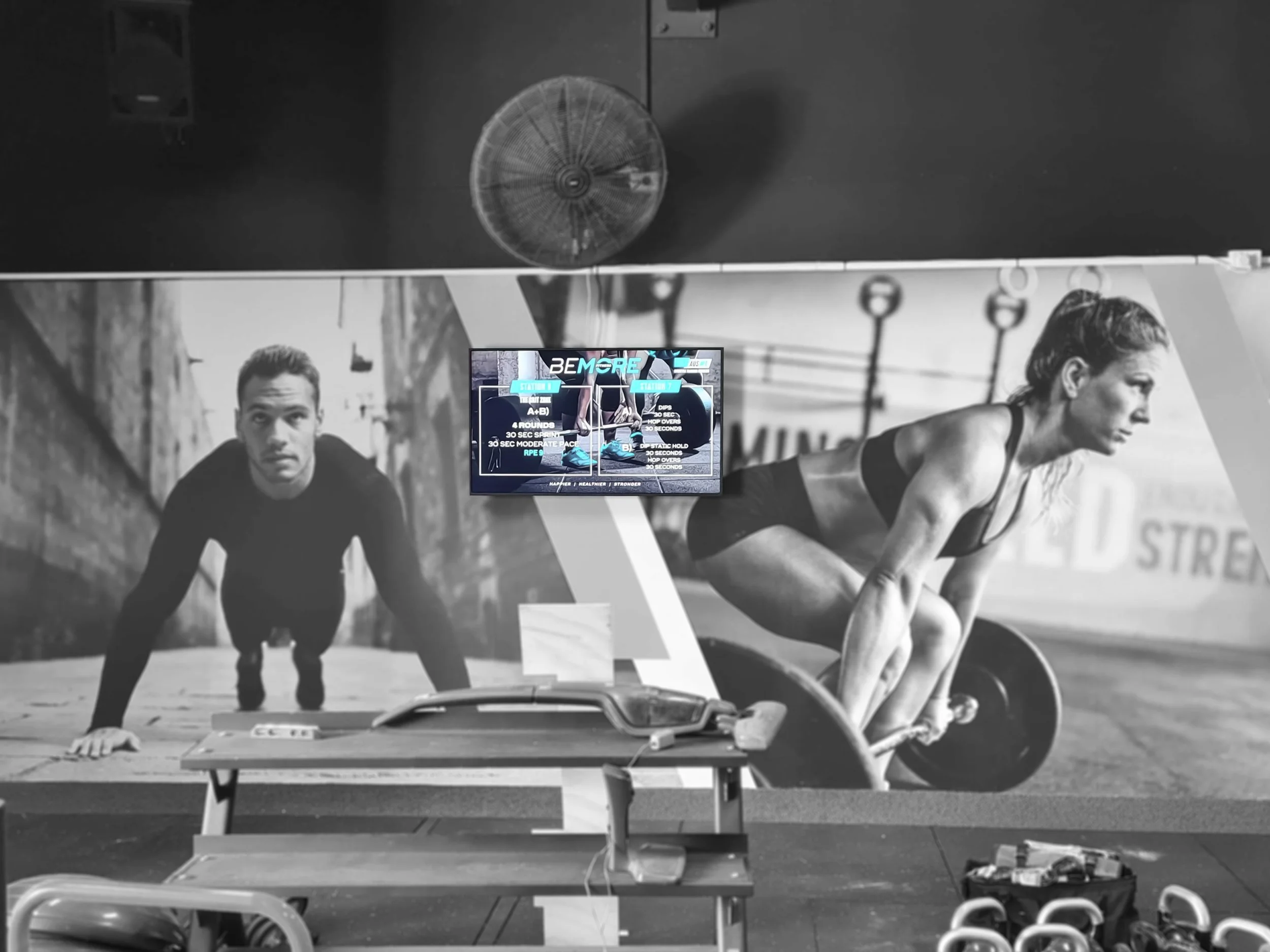 Wall-mounted gym display showing two workout stations with interval times and rounds at Bemore Fitness.