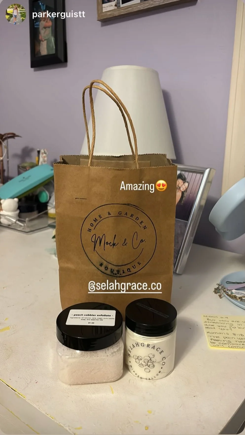 A paper shopping bag from Mock & Co. Home & Garden on a white table, two containers of bath salts or body scrub, and some jewelry. A photo frame and other items are on a purple wall behind the table.