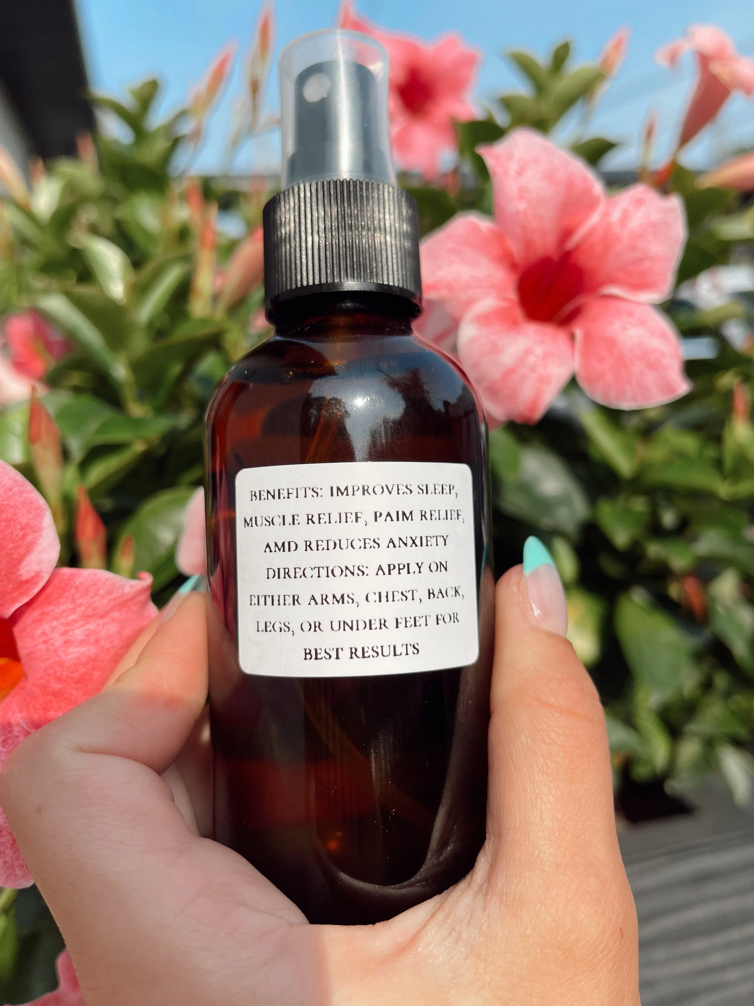 A hand holding a dark amber spray bottle with a label containing benefits and directions, in front of pink flowers and green foliage.
