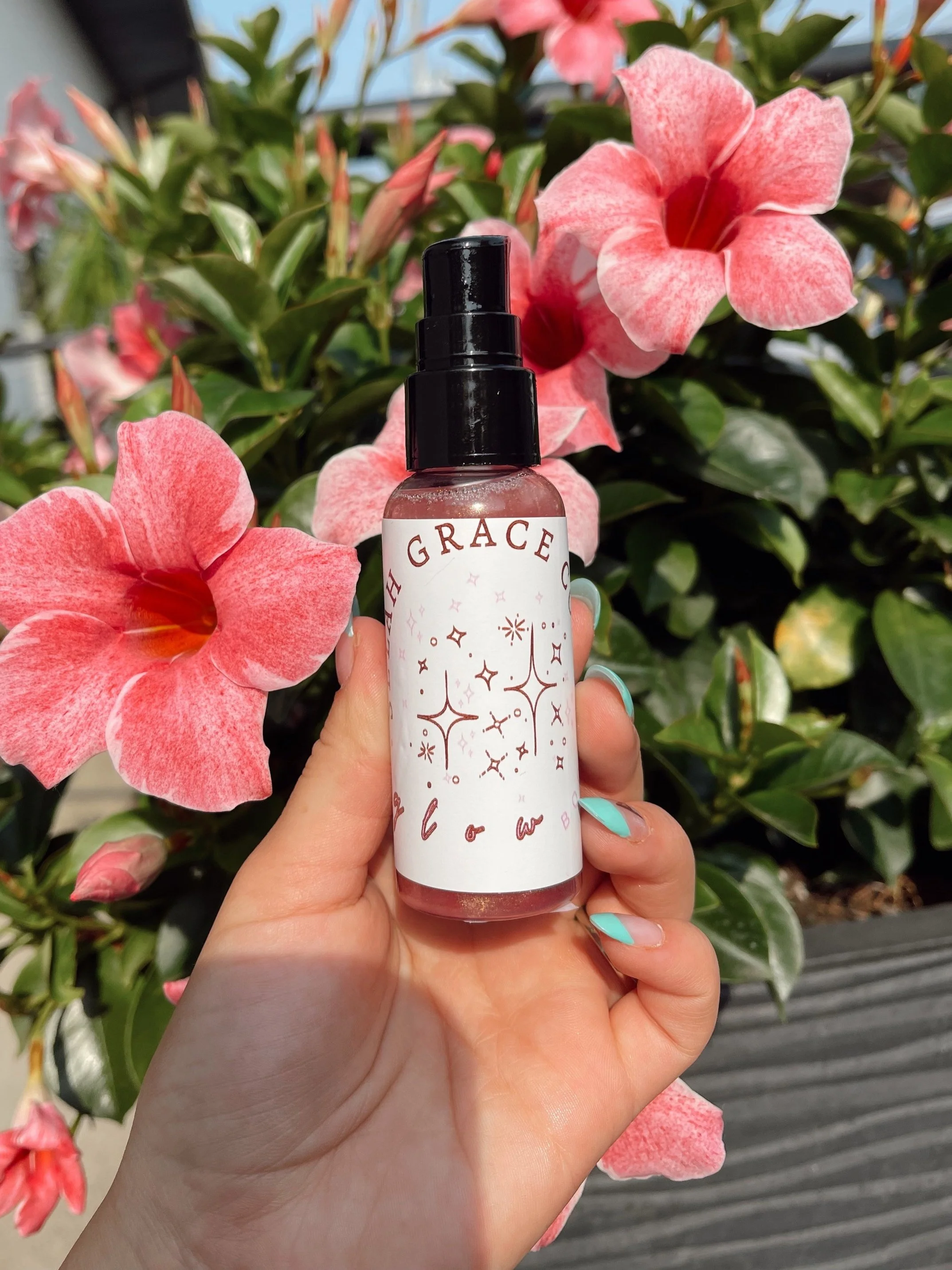 A hand holding a spray bottle labeled 'High Grace' with a design of stars and sparkles, with pink and red flowers and green leaves in the background.
