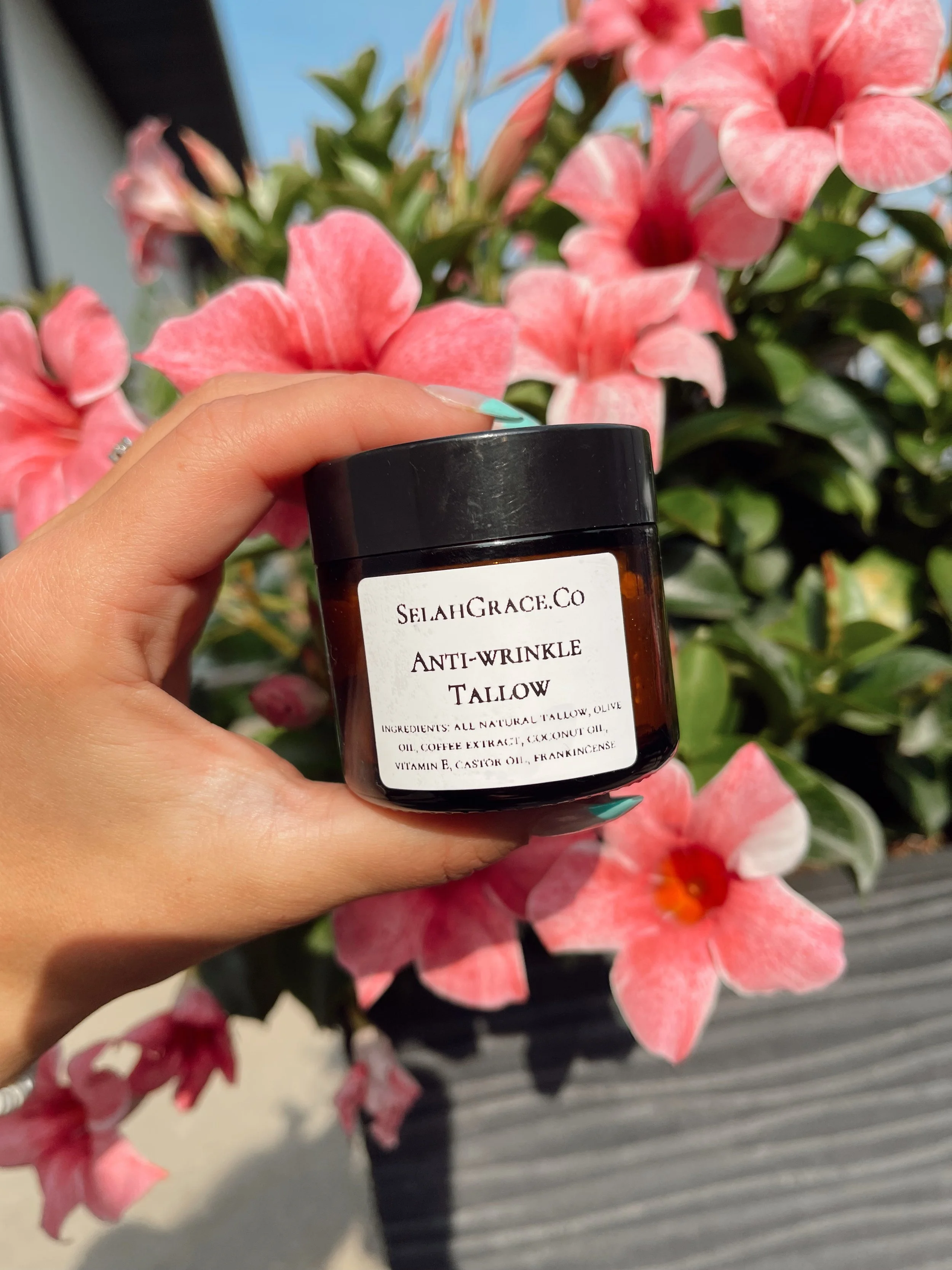A hand holding a jar of Selah Grace Co Anti-Wrinkle Tallow in front of pink and white flowers outdoors.