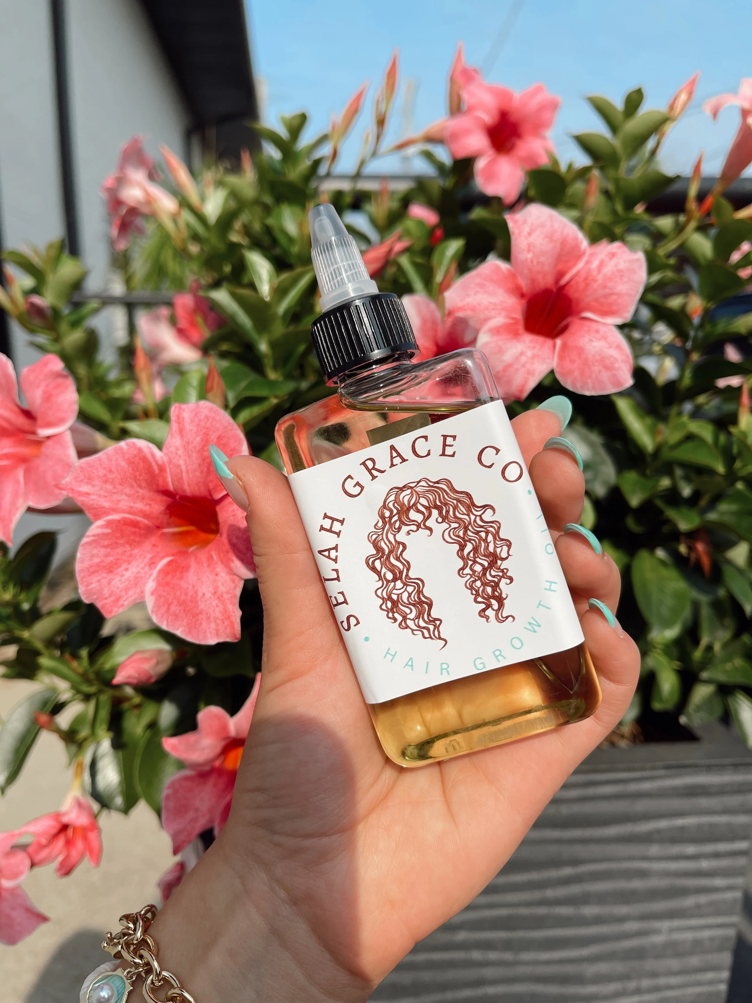 A hand holding a bottle of hair growth oil in front of pink and peach flowers in bloom, with clear sky and a building in the background.