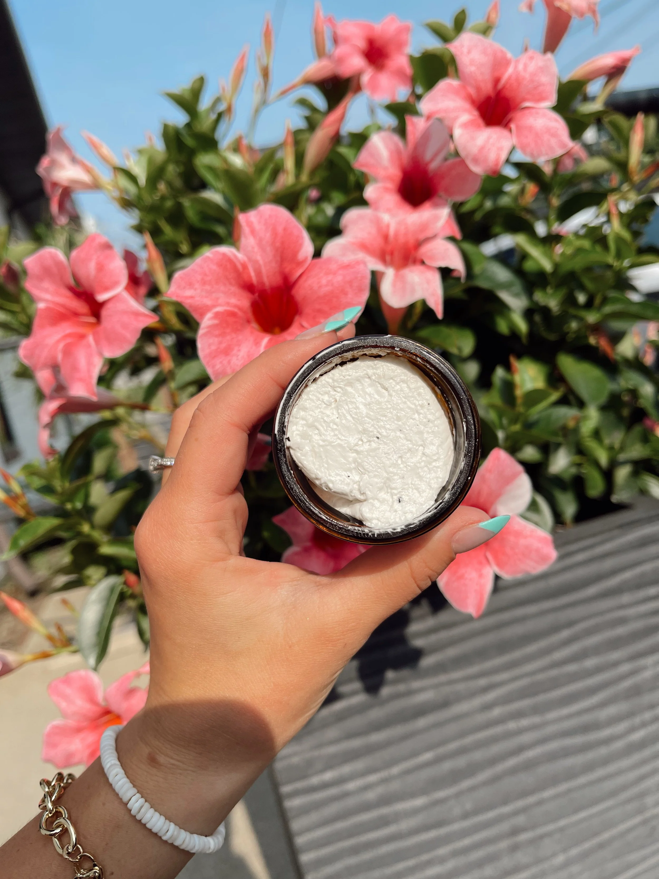 Hand holding open jar of white cream or lotion with pink flowering plants in the background.