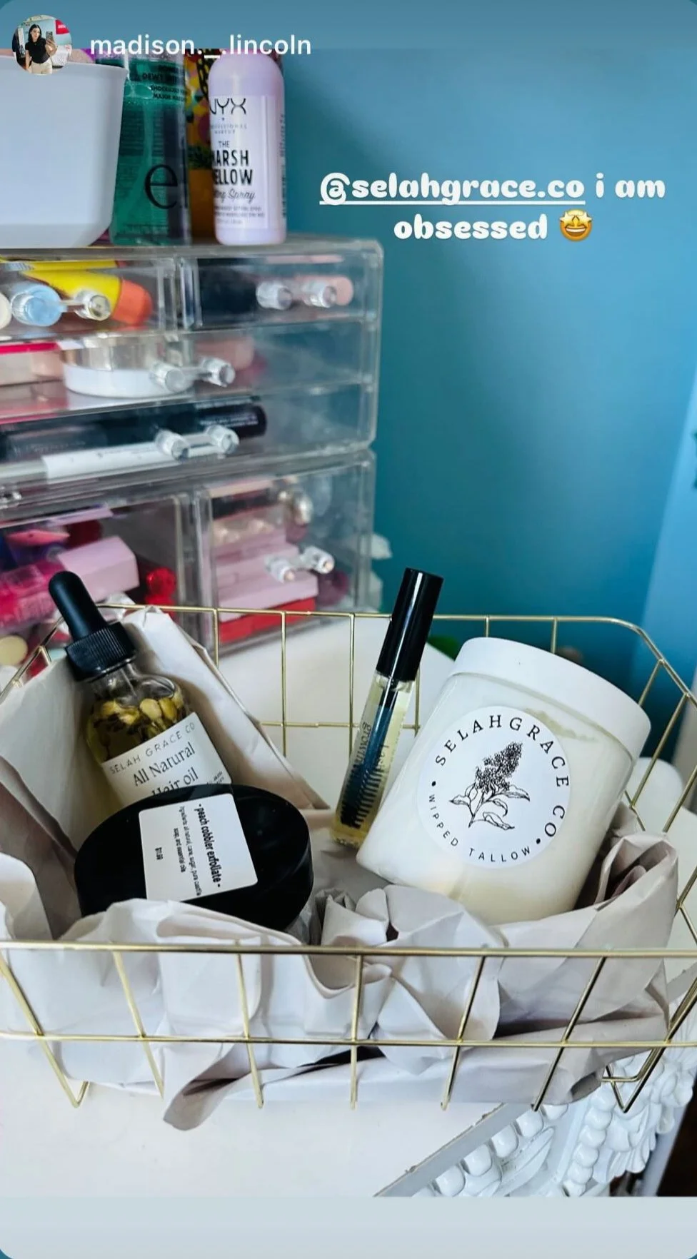 A gold wire basket containing skincare and beauty products, including a candle labeled 'Selah Grace,' a rollerball oil, a black container, a small bottle with yellowish oil and herbs, and a tester vial of liquid. In the background is a clear acrylic drawer organizer with various small bottles and containers, against a blue wall.