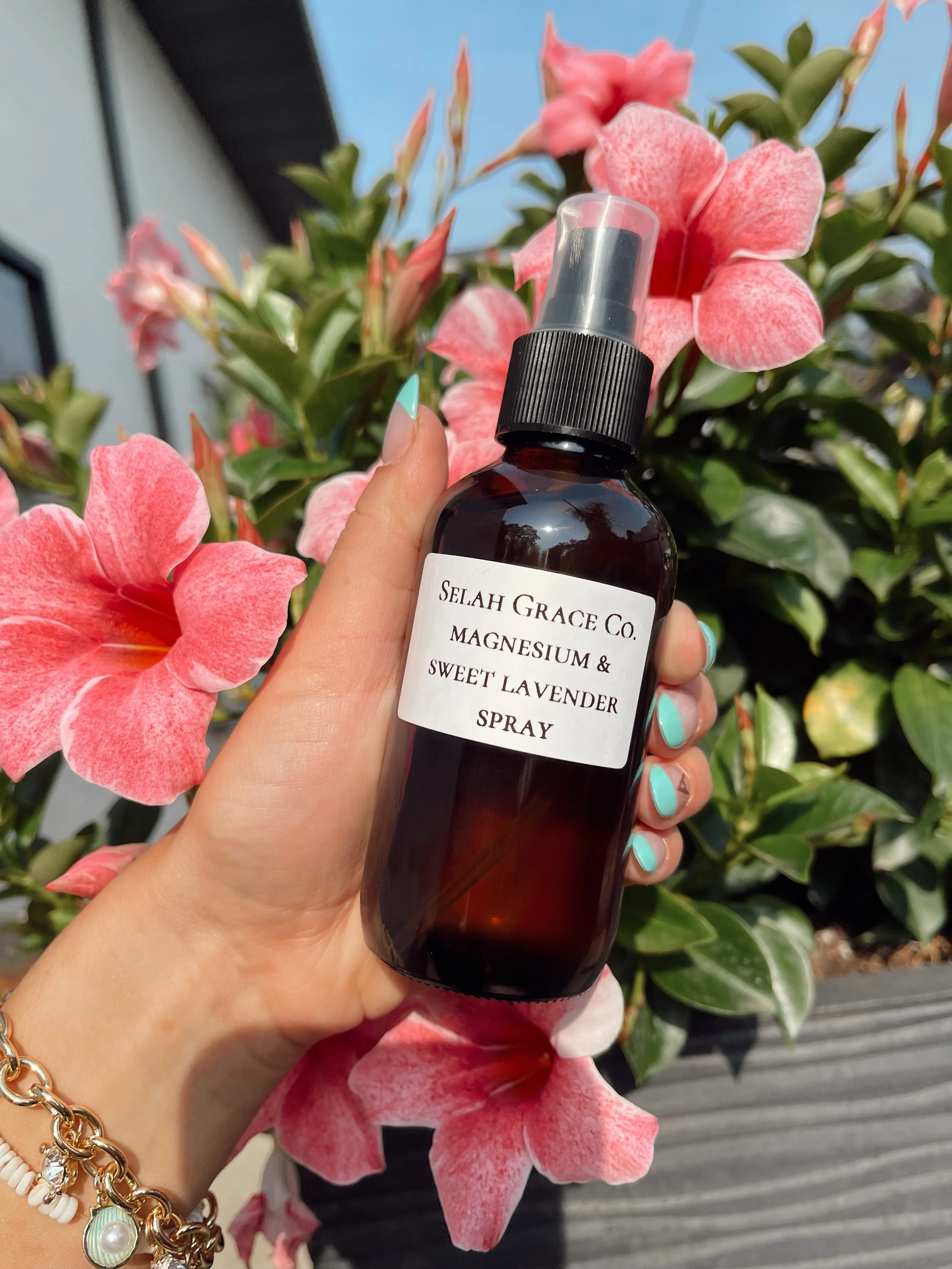 A hand holding a brown spray bottle labeled 'Selah Grace Co Magnesium & Sweet Lavender Spray' in front of pink flowers and green leaves outdoors.