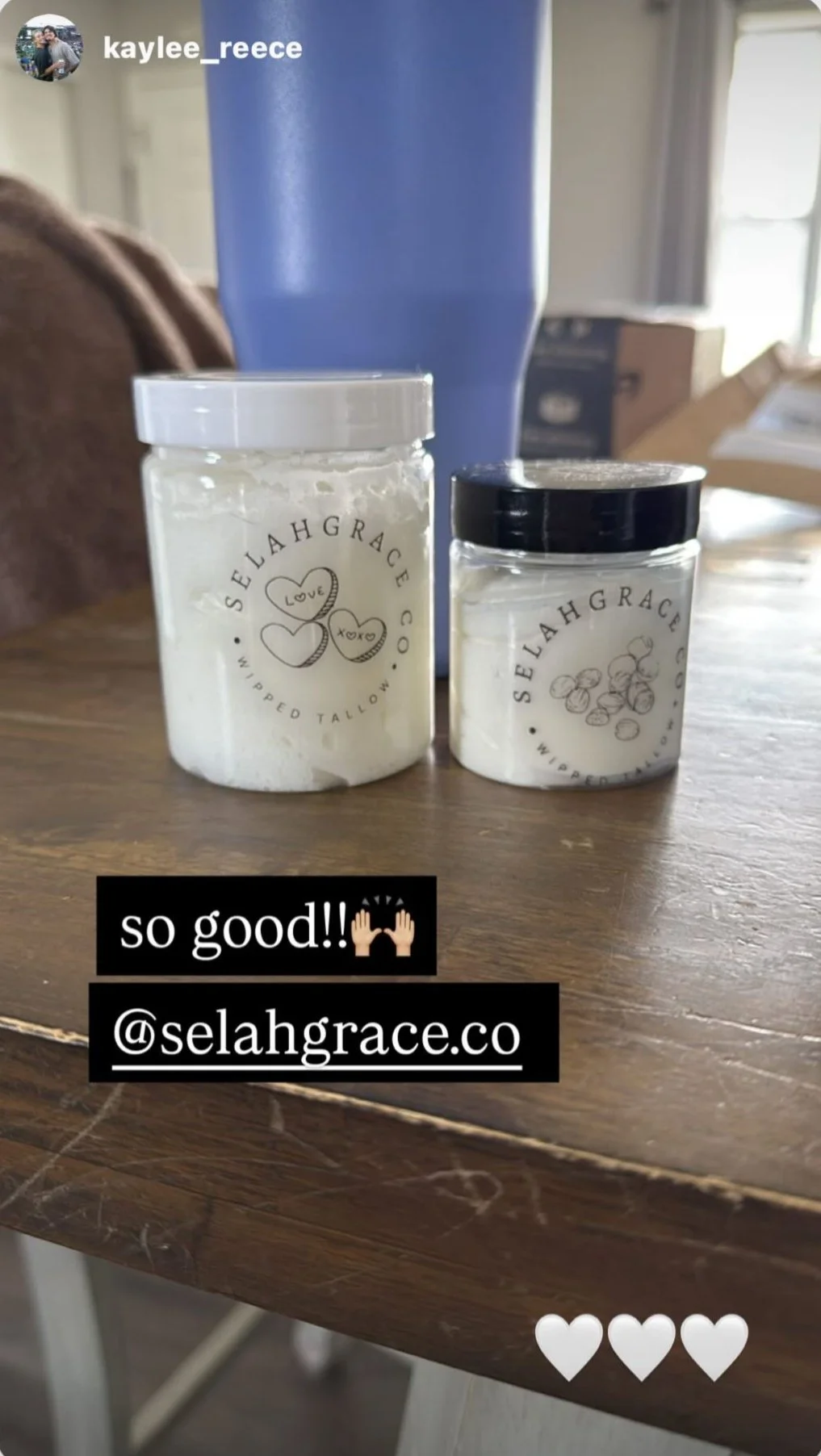 Two jars of whipped body butter on a wooden table, one jar with a white lid and the other with a black lid, from Selah Grace Co, with a blue cup and boxes in the background.