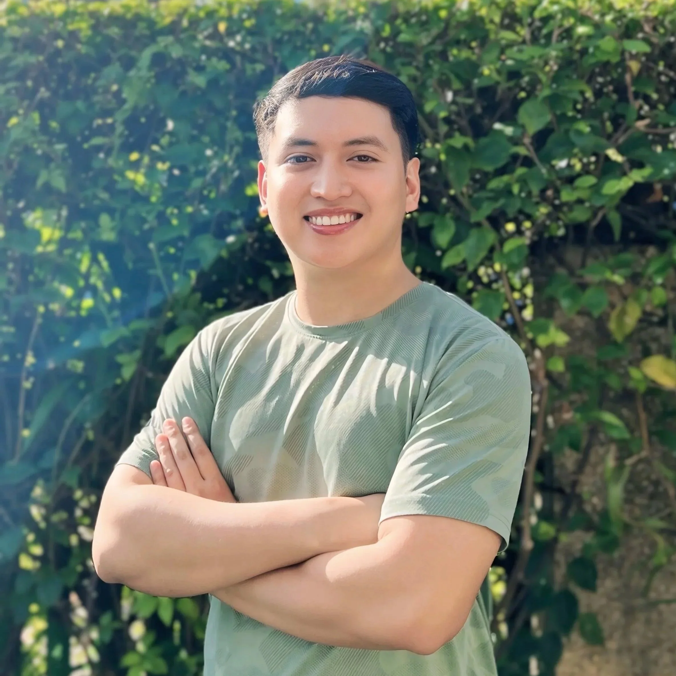 Young male dietitian with short dark hair smiling, crossing arms, outdoors in front of green leafy background, wearing a light green shirt.