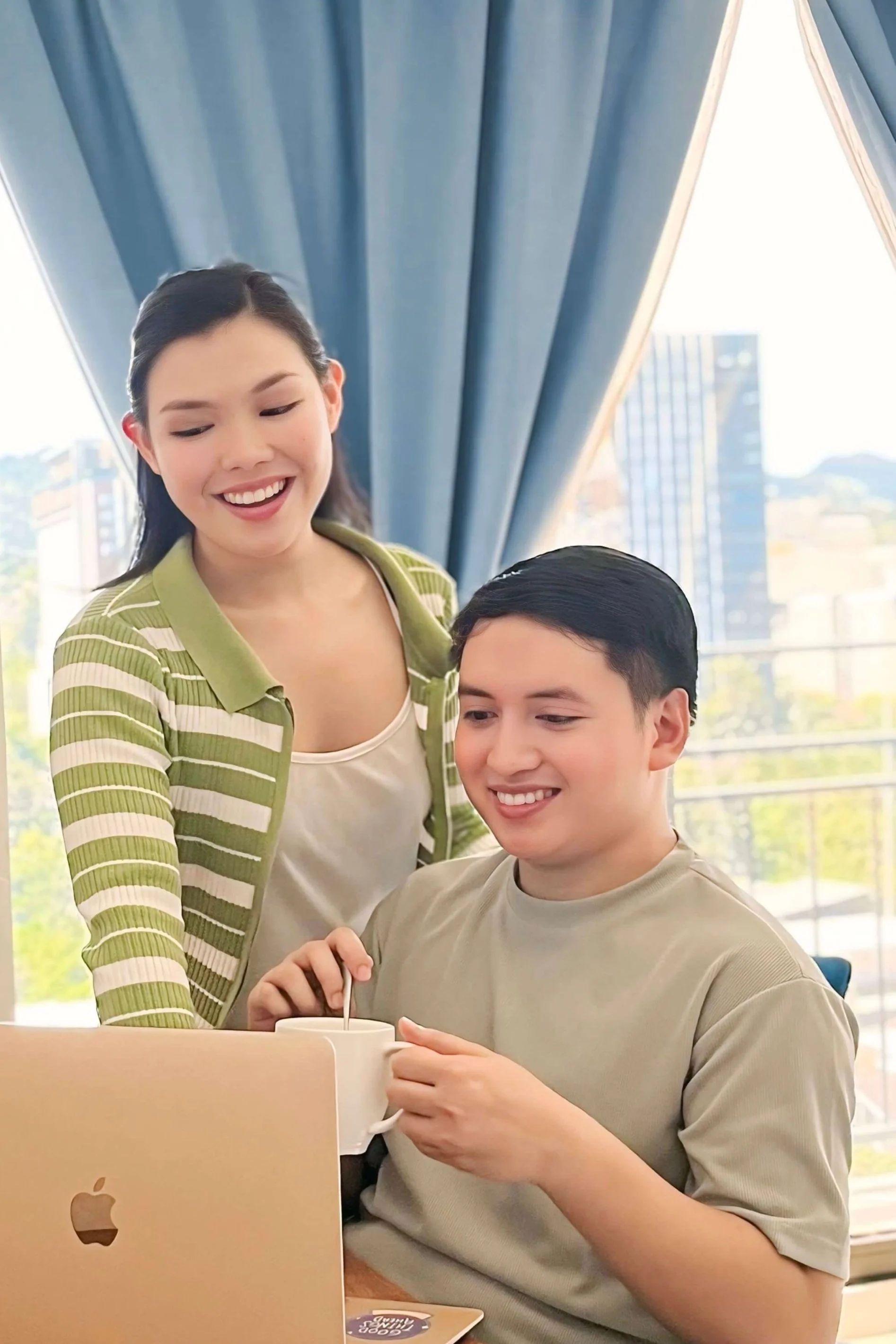 Two dietitians consulting a patient at a desk using a laptop, both smiling. The man holds a cup and stirs with a spoon. They are near a window with blue curtains and a view of buildings outside.
