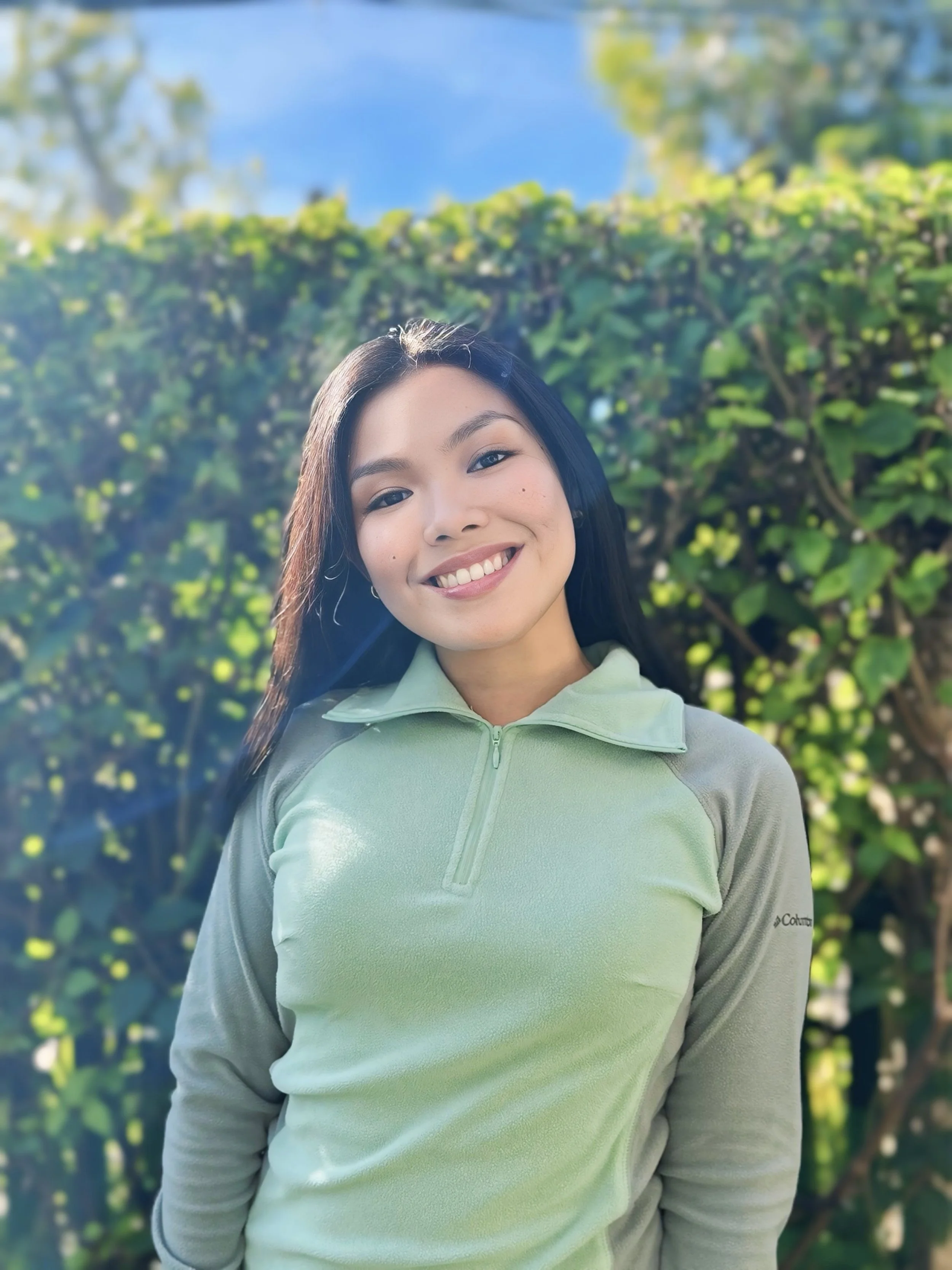Dietitian smiling outdoors wearing a light green long-sleeve top, standing in front of a leafy green hedgerow with a blue sky in the background.