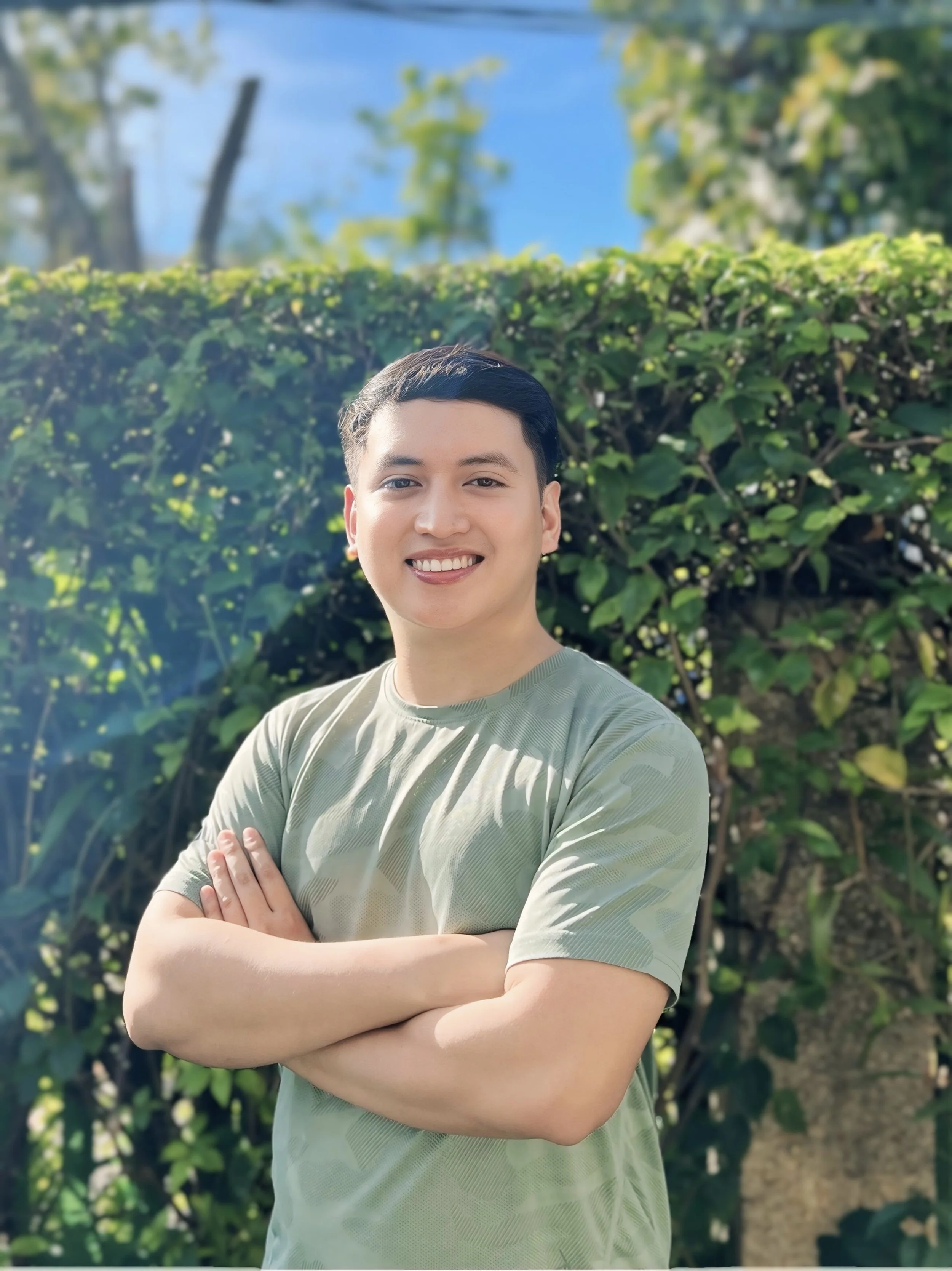 Dietitian smiling and standing outdoors with arms crossed, wearing a light green shirt, in front of a hedge, under a clear blue sky.