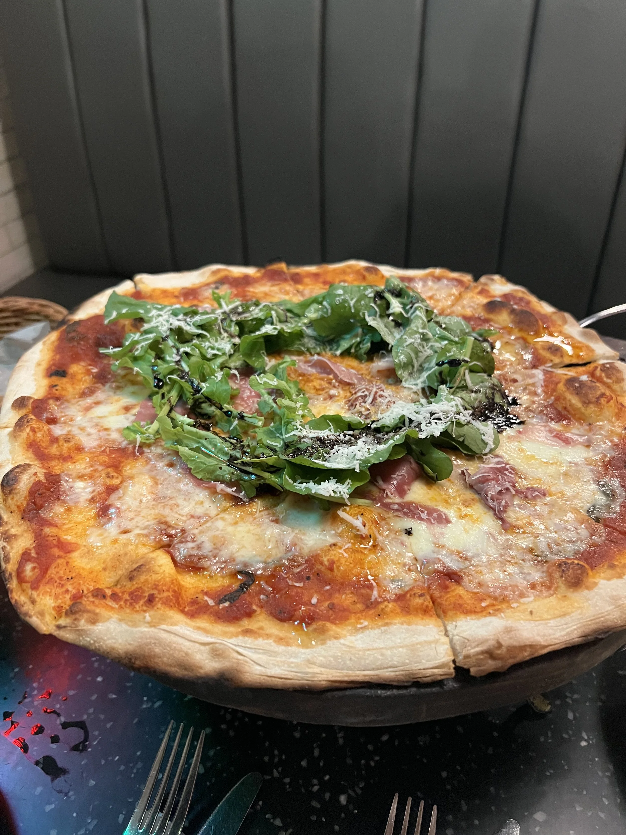 A pizza atop a black speckled table, topped with fresh greens, cheese, tomato sauce, and various meats. Forks and knives are placed on the table around the pizza.