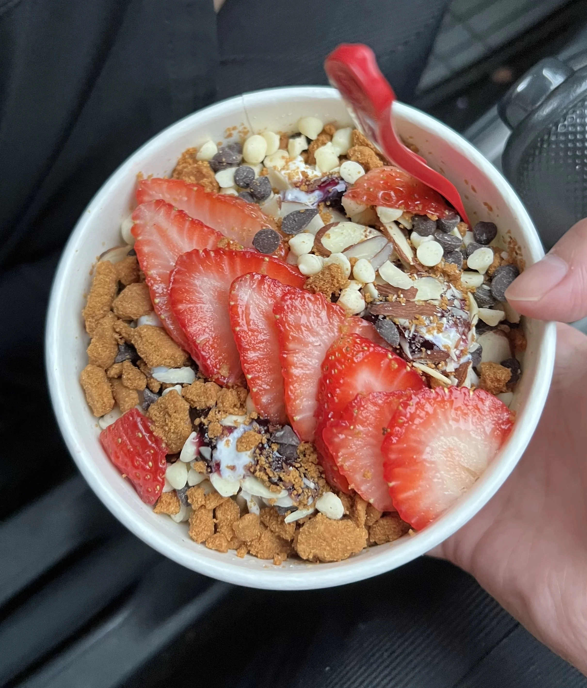 A close-up of a cup of yogurt topped with sliced strawberries, white chocolate chips, and crushed cookies, held by a person's hand.