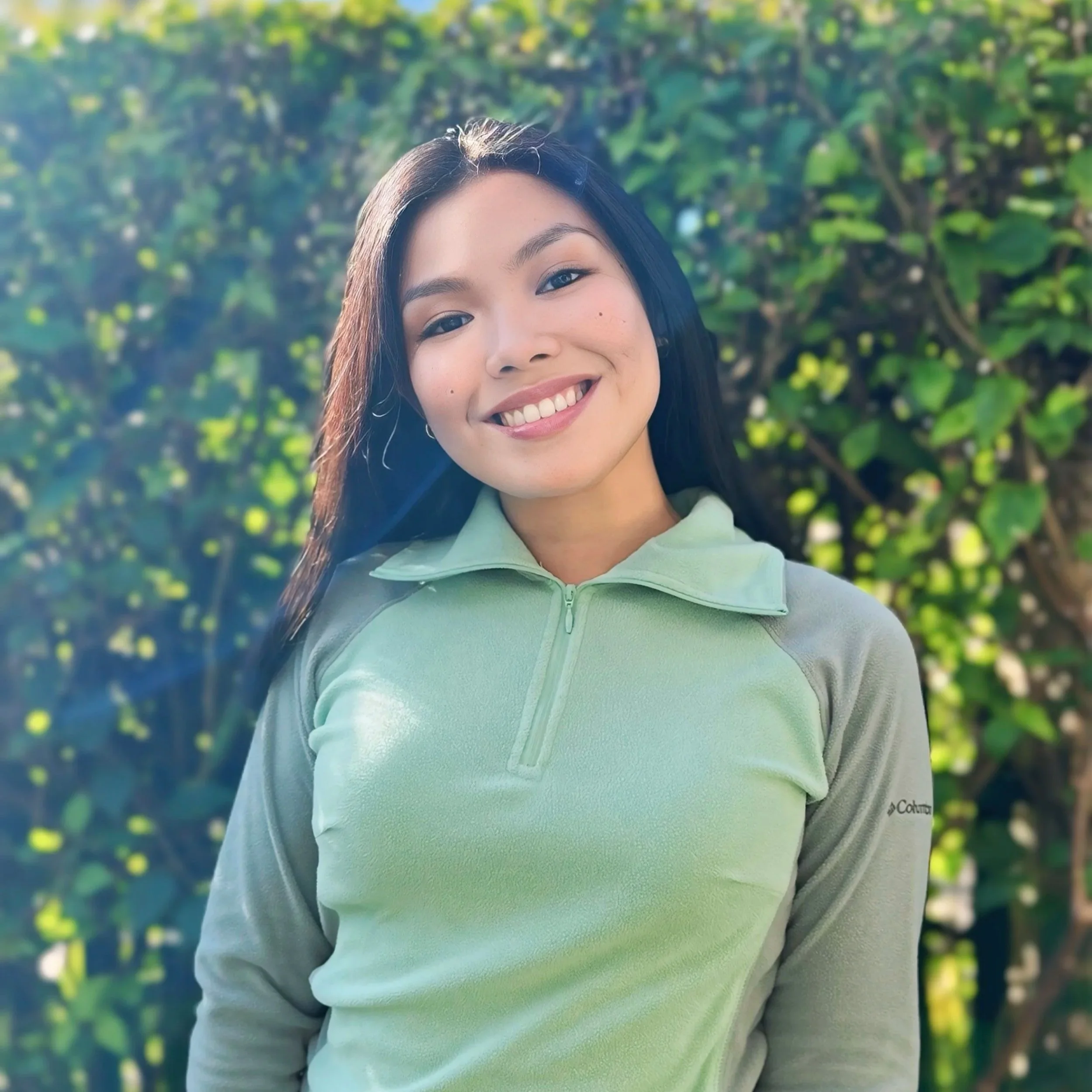 A young female dietitian smiling outdoors, wearing a light green zip-up jacket, with greenery in the background.